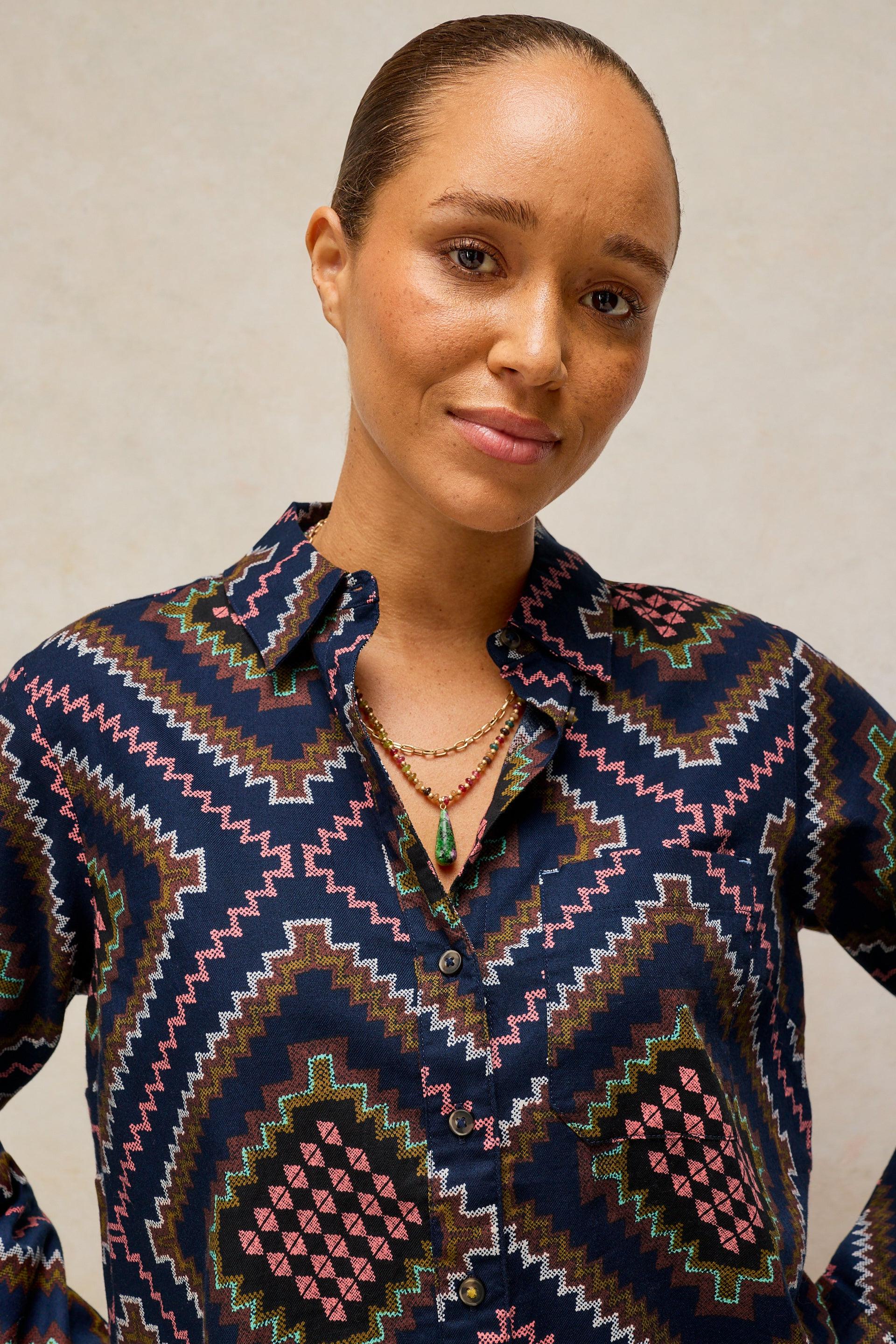 A woman wearing a long-sleeve button-up shirt with a colorful geometric pattern in navy, pink, brown, and white, paired with layered gold necklaces, set against a neutral beige background.