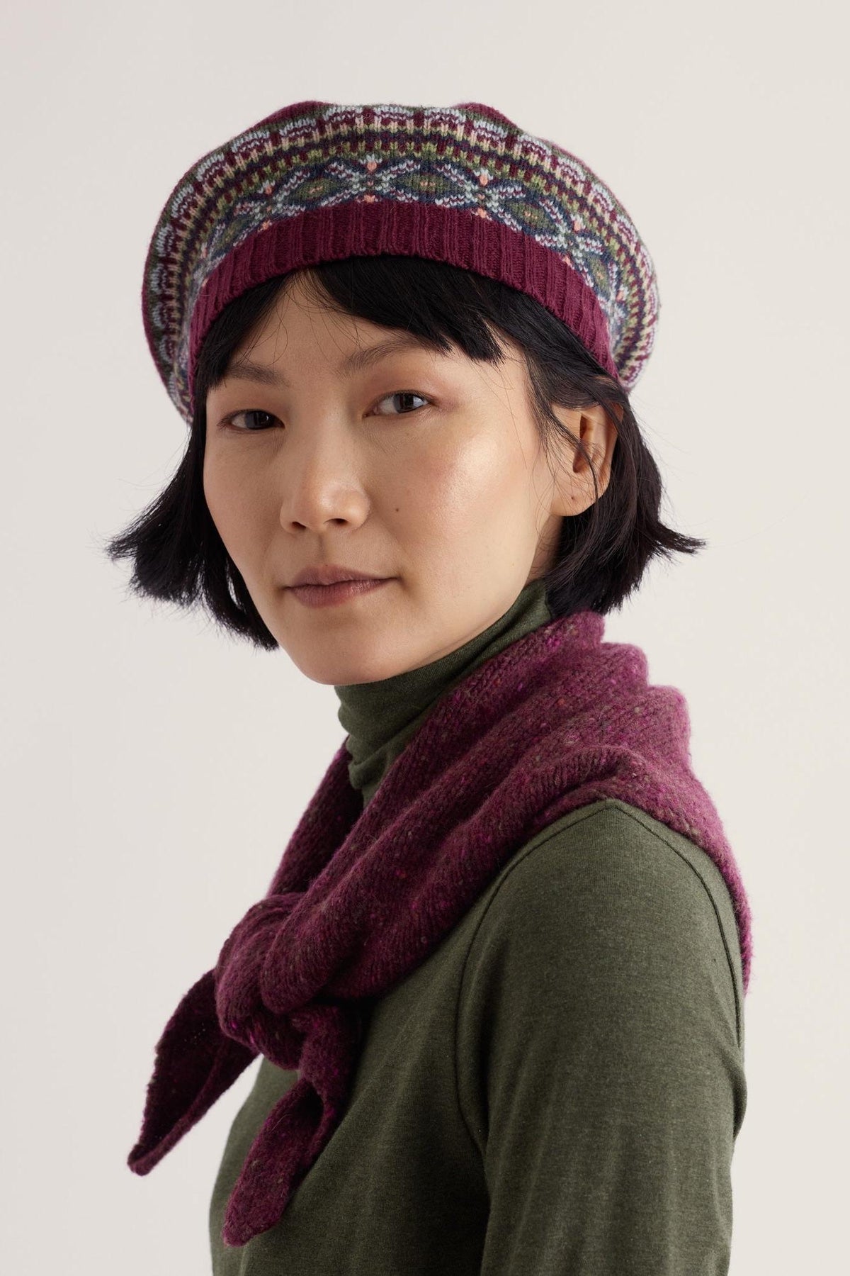 Woman with dark hair wearing a patterned Seasalt Cornwall wool beret and a dark green top with a purple scarf.