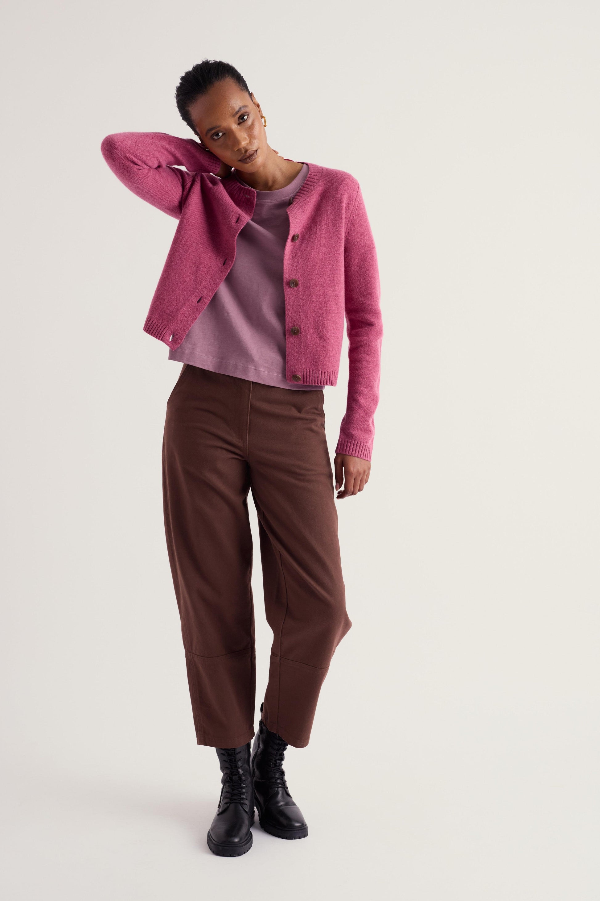 A model posing in a studio setting wearing a pink cropped cardigan over a mauve top, brown cropped trousers, and black boots against a light gray background.