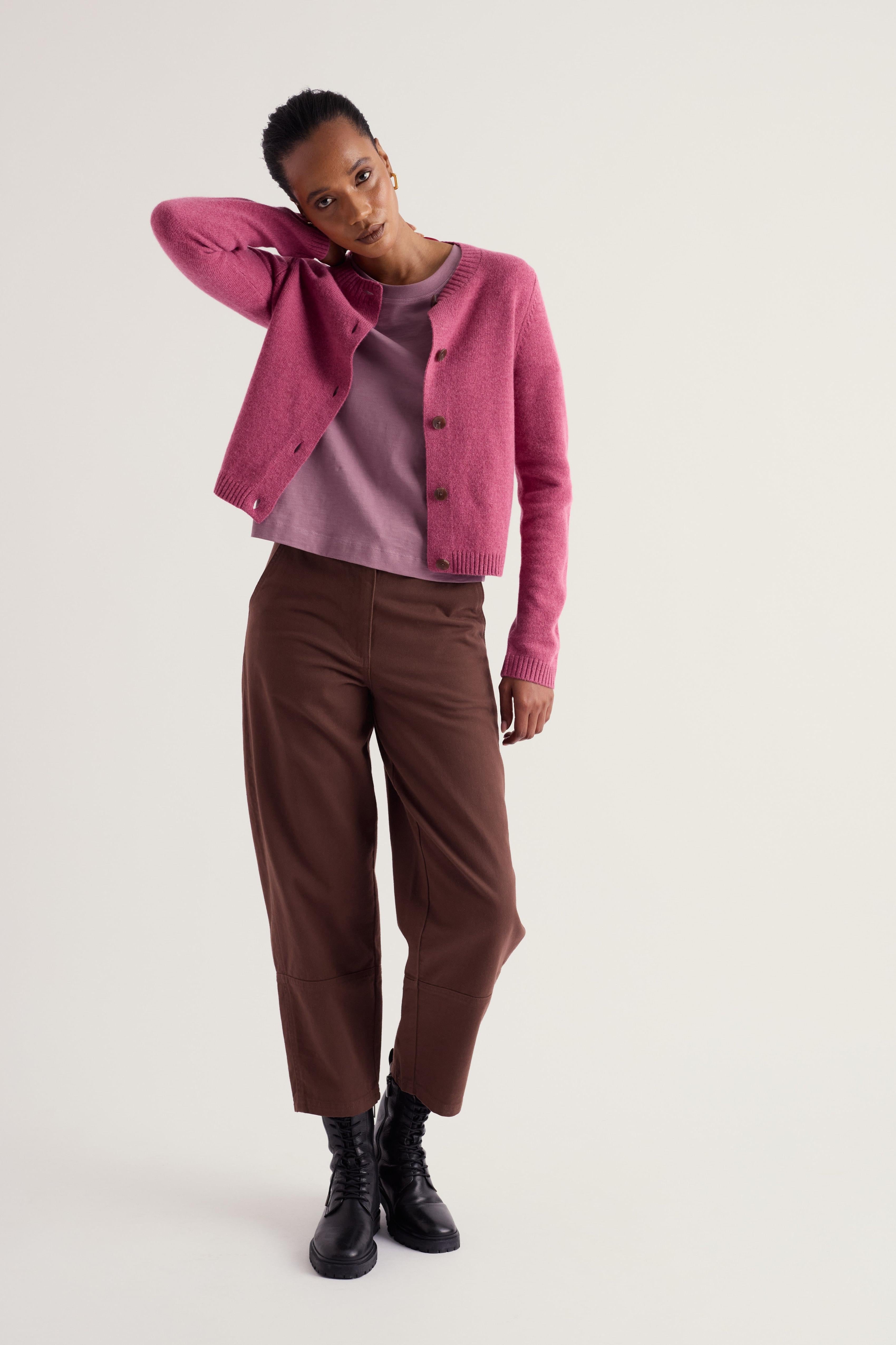 A model posing in a studio setting wearing a pink cropped cardigan over a mauve top, brown cropped trousers, and black boots against a light gray background.