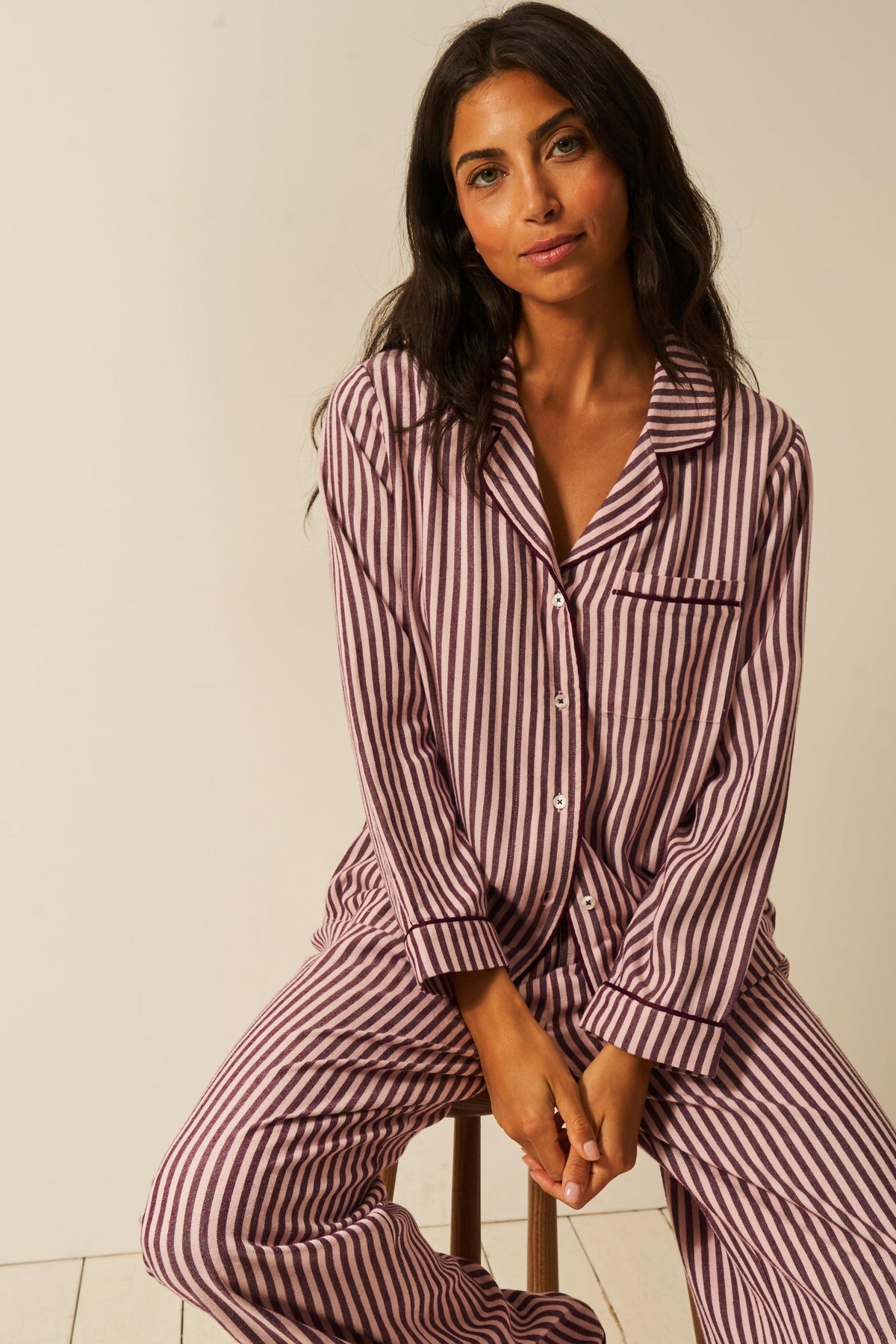 A woman wearing soft pink stripe tencel pyjamas sitting on a stool against a plain background.
