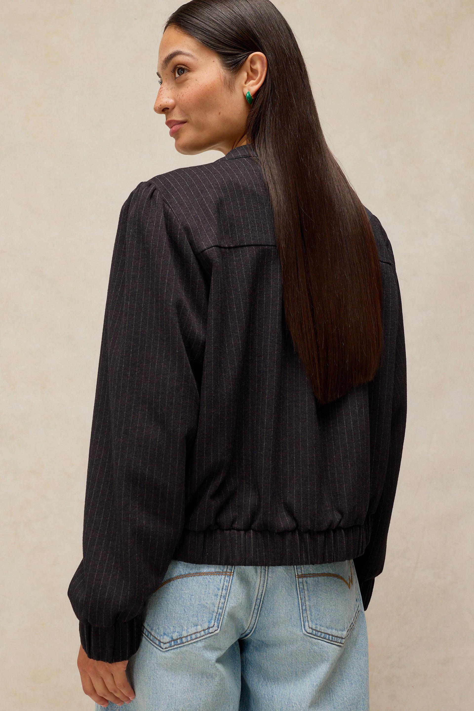 A woman with long dark brown hair is shown from the back wearing a navy pinstriped bomber jacket and light blue jeans, set against a textured beige background.