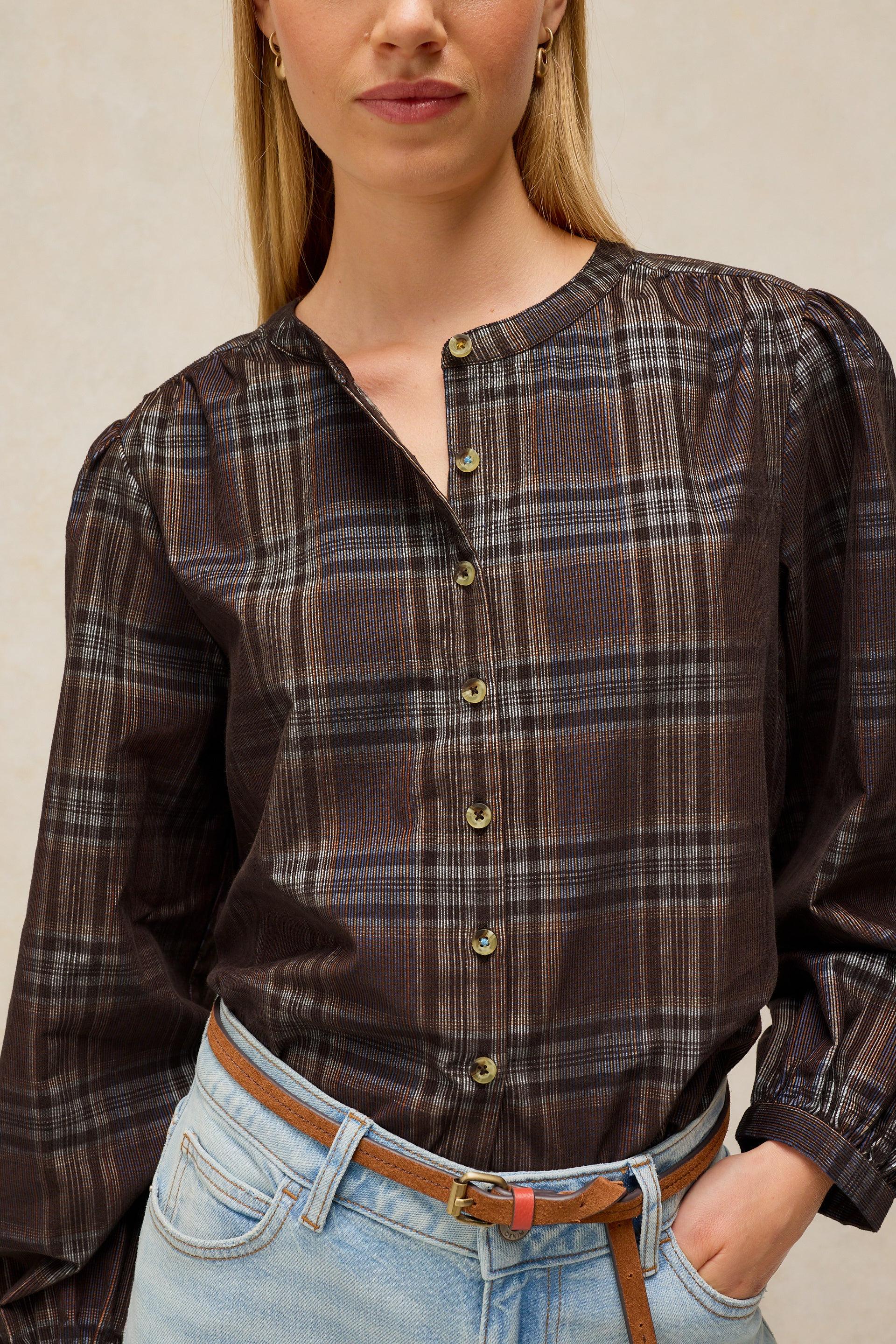 A woman wearing a dark brown plaid button-up blouse with puff sleeves and gold-tone buttons, paired with light-wash blue jeans and a brown belt.
