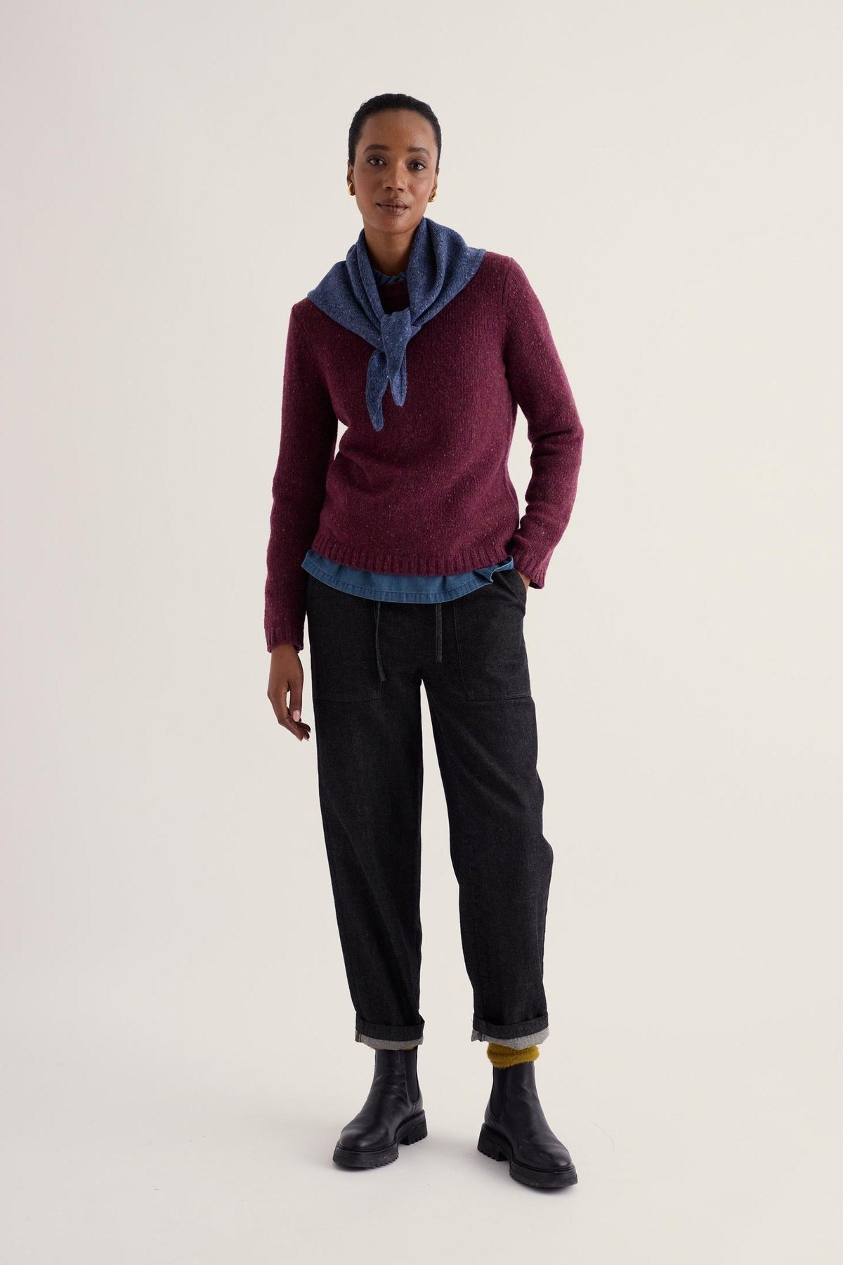 A woman modeling a plum merino wool jumper and blue scarf from Seasalt Cornwall against a neutral background.