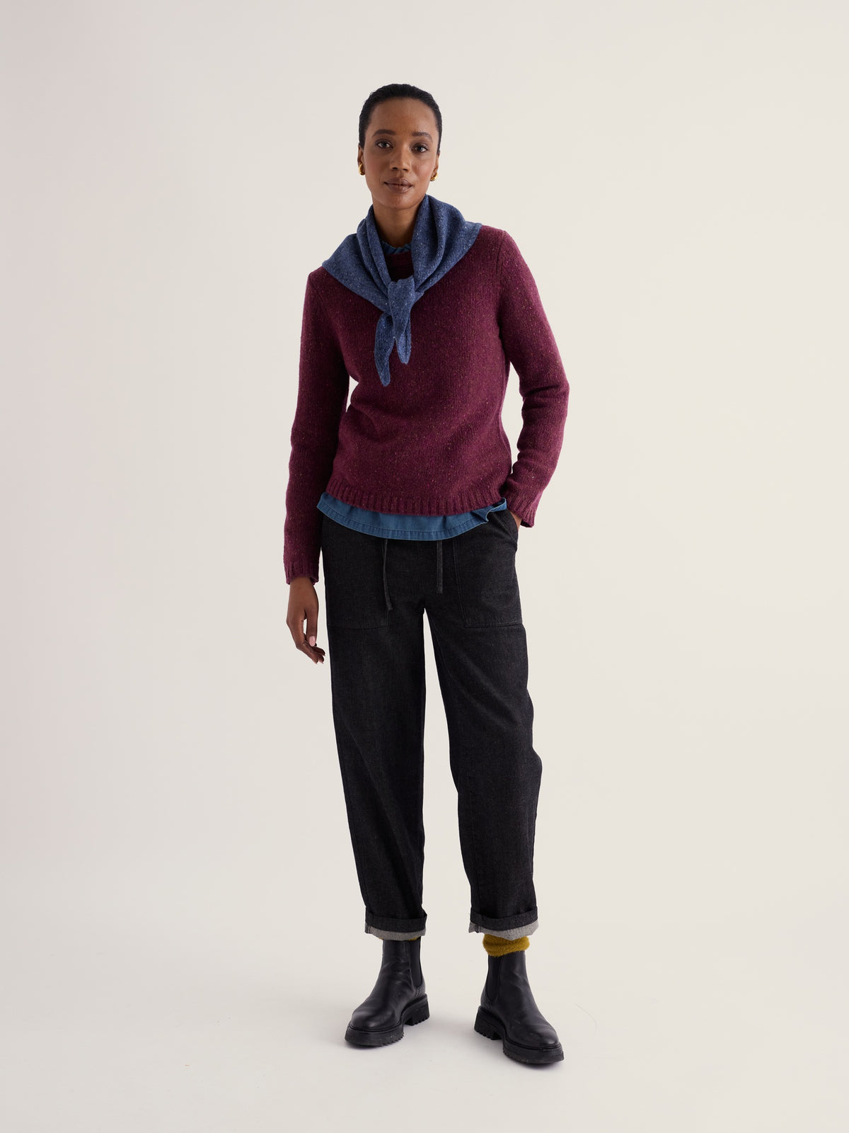 A woman modeling a plum merino wool jumper and blue scarf from Seasalt Cornwall against a neutral background.