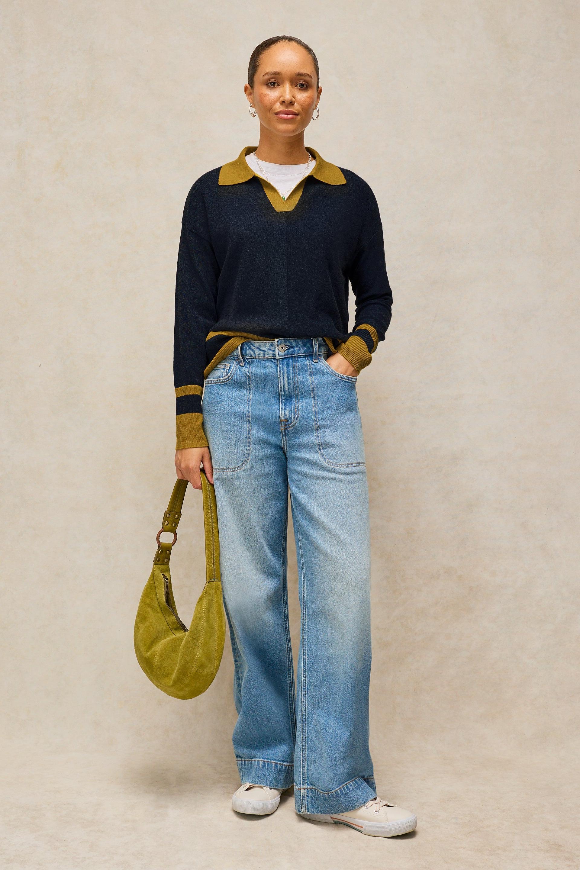 A model stands against a textured beige studio backdrop wearing a navy long-sleeve knit sweater with mustard yellow collar and cuffs, light blue wide-leg jeans, white sneakers, and holding a mustard yellow crescent shoulder bag.