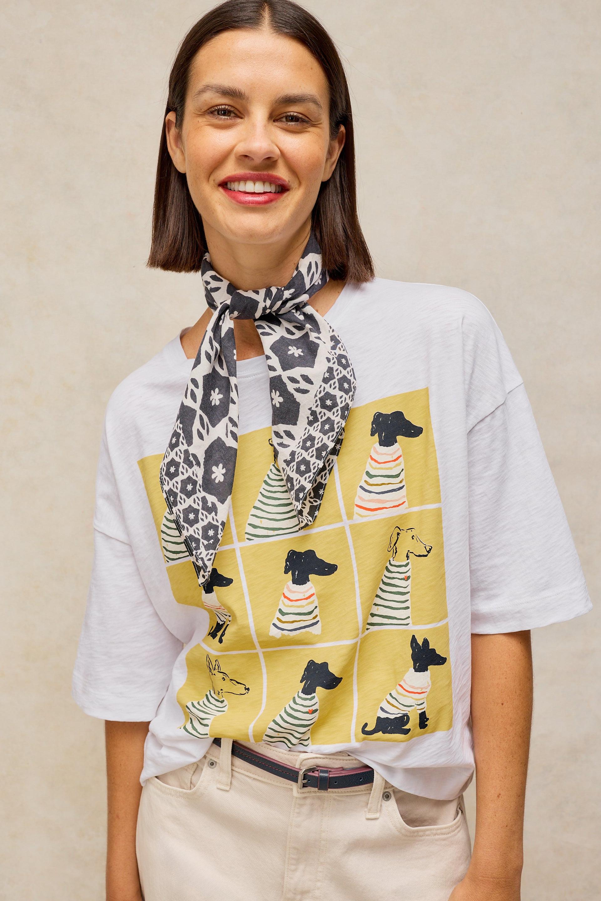 A woman wearing a white graphic t-shirt with a yellow dog print, a black and white patterned scarf tied around her neck, and beige pants, set against a neutral beige background.