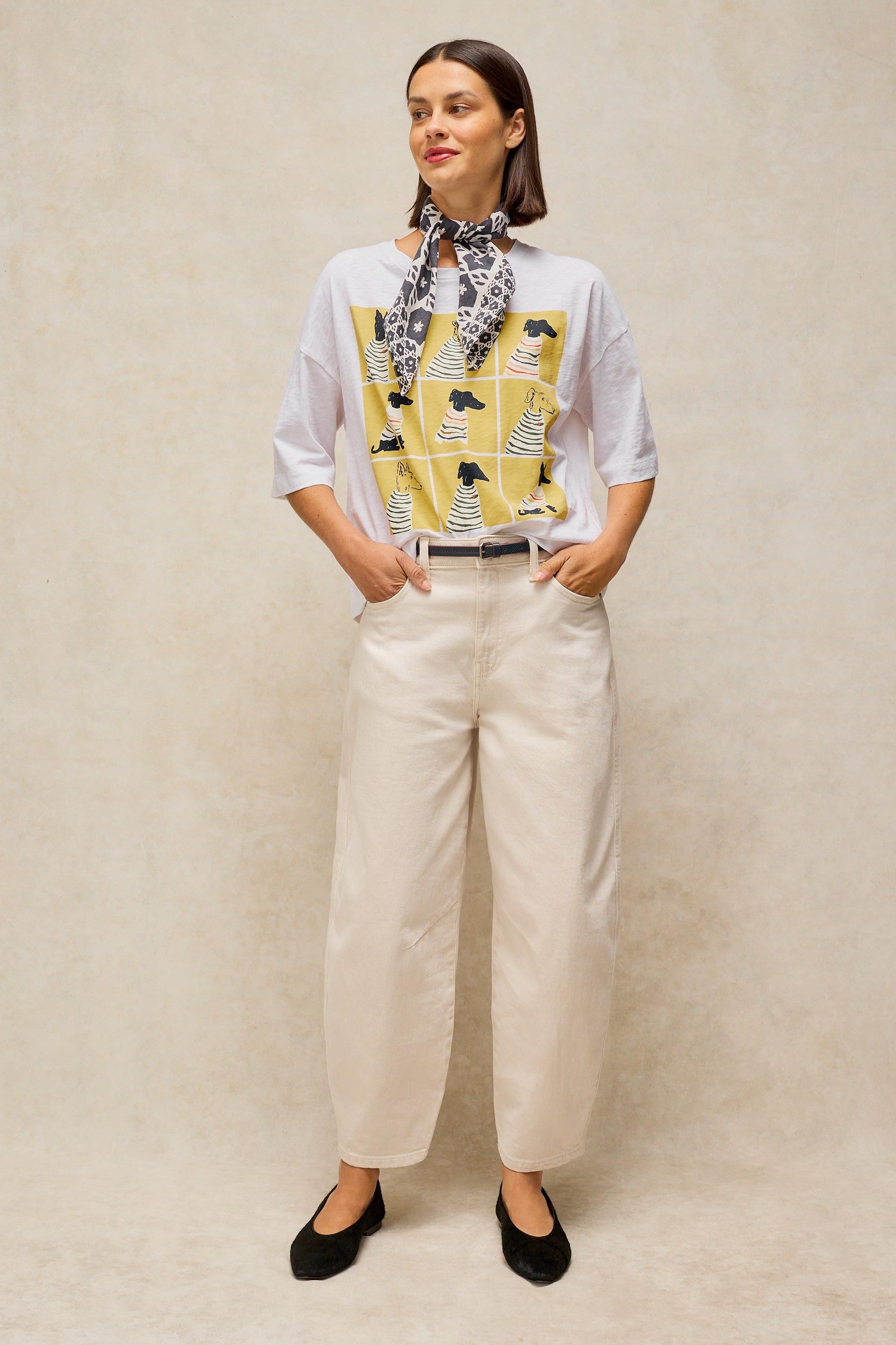 A woman modeling a white short-sleeve top with a yellow and black graphic print on the front, paired with cream wide-leg trousers and black flats, standing against a textured beige studio backdrop.