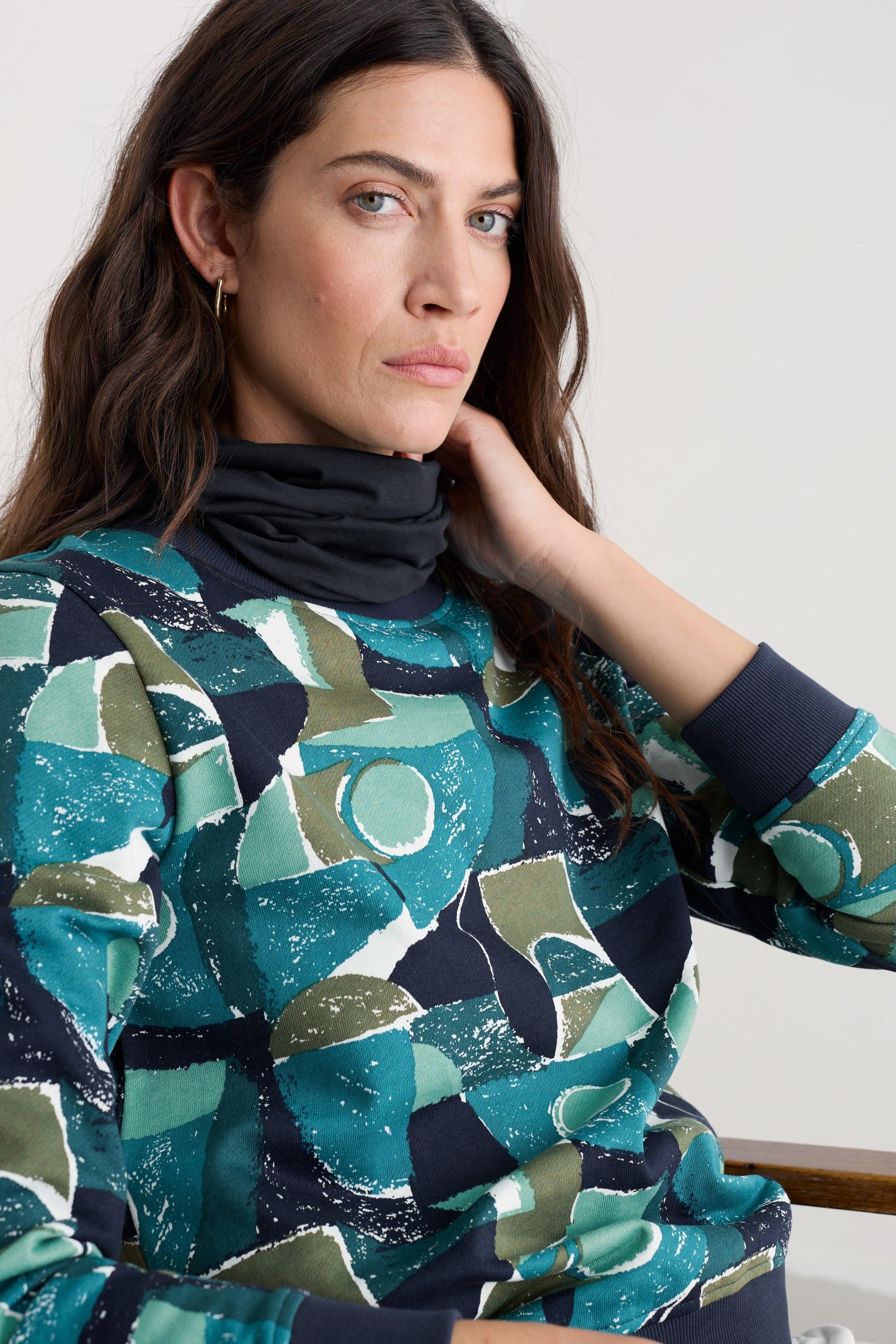 A woman wearing a long-sleeve crewneck sweatshirt with an abstract geometric pattern in teal, green, navy, and white, paired with a black infinity scarf, set against a plain light gray background.