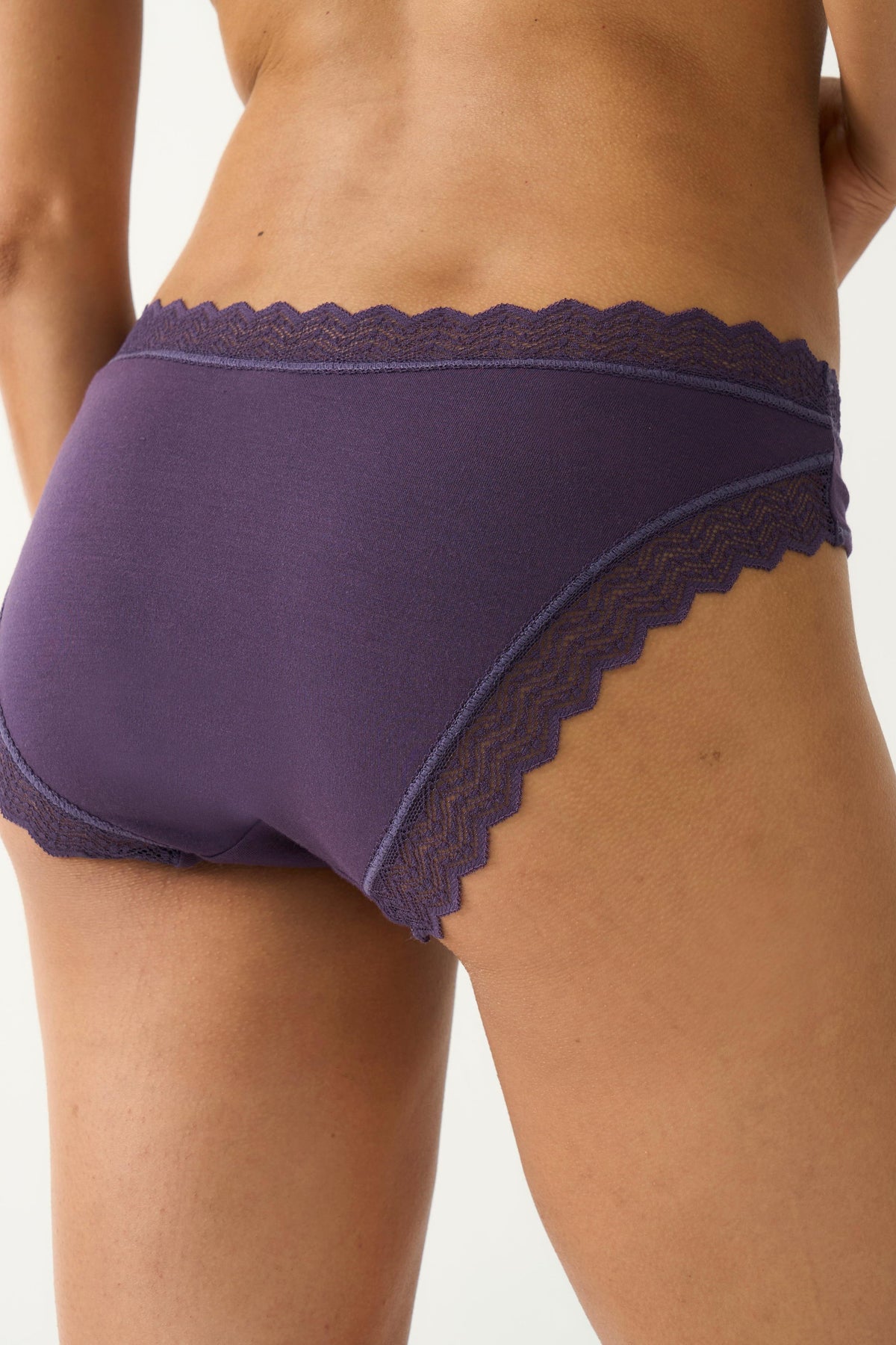 Rear view of a woman wearing purple classic cut underwear with lace trim against a plain white background.