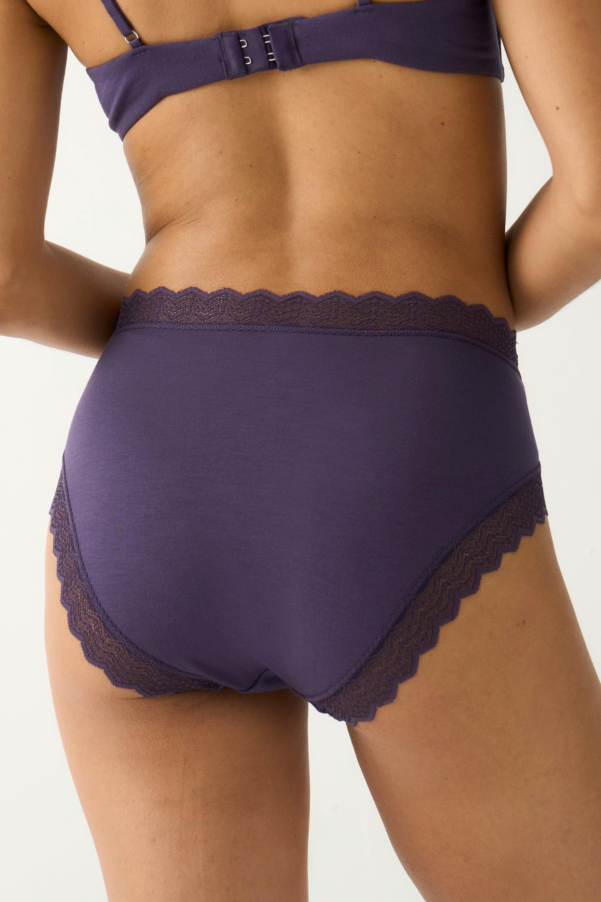 Rear view of a woman wearing purple high-rise underwear with lace trim against a white background.
