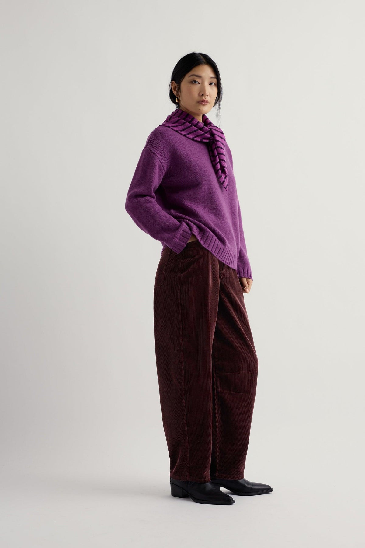 Woman wearing a purple knit jumper and scarf with dark corduroy trousers against a plain white background.