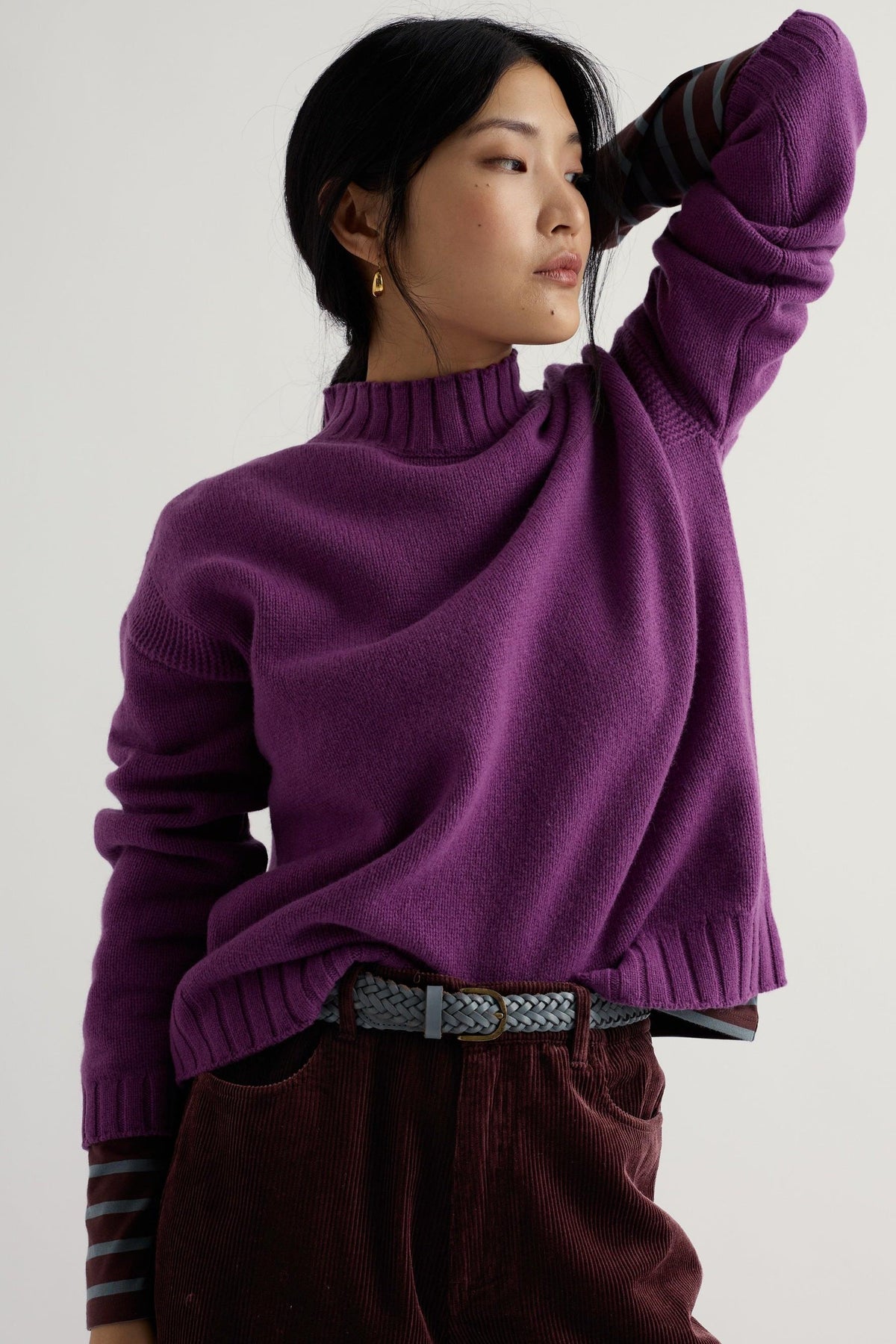 A woman posing in a vibrant purple knit sweater and corduroy trousers against a plain studio background.