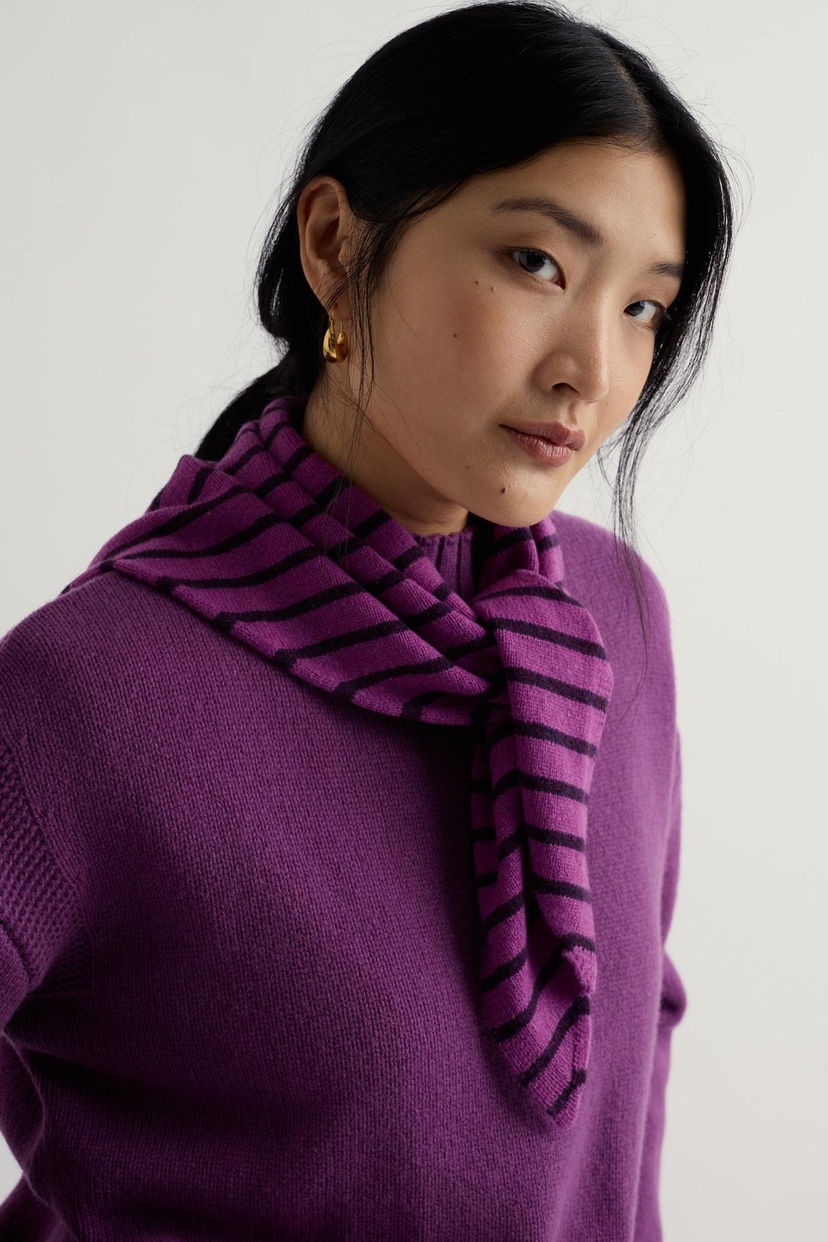 A woman wearing a purple lambswool knit sweater with a matching striped scarf against a plain background.