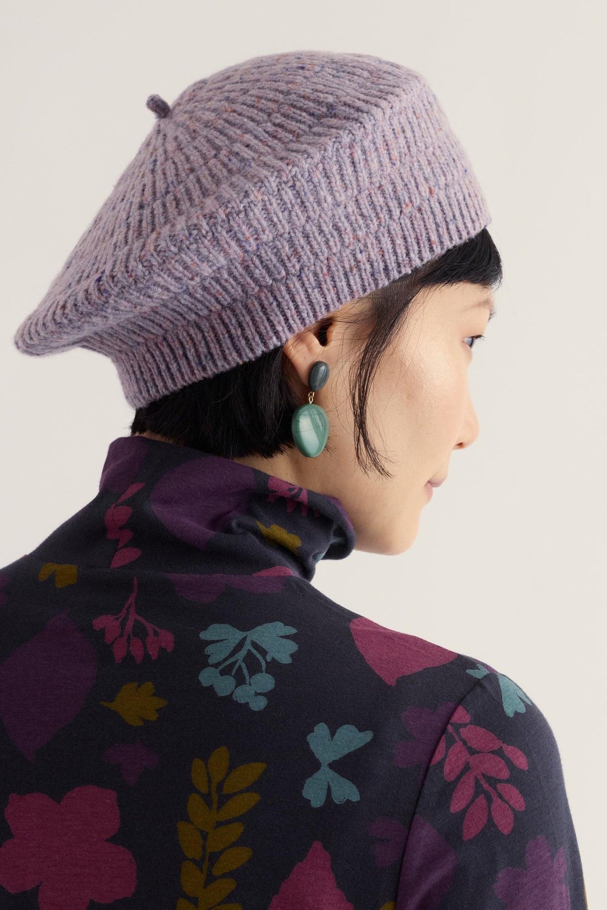 Woman wearing a purple knitted merino wool beret hat and patterned turtleneck against a neutral background.