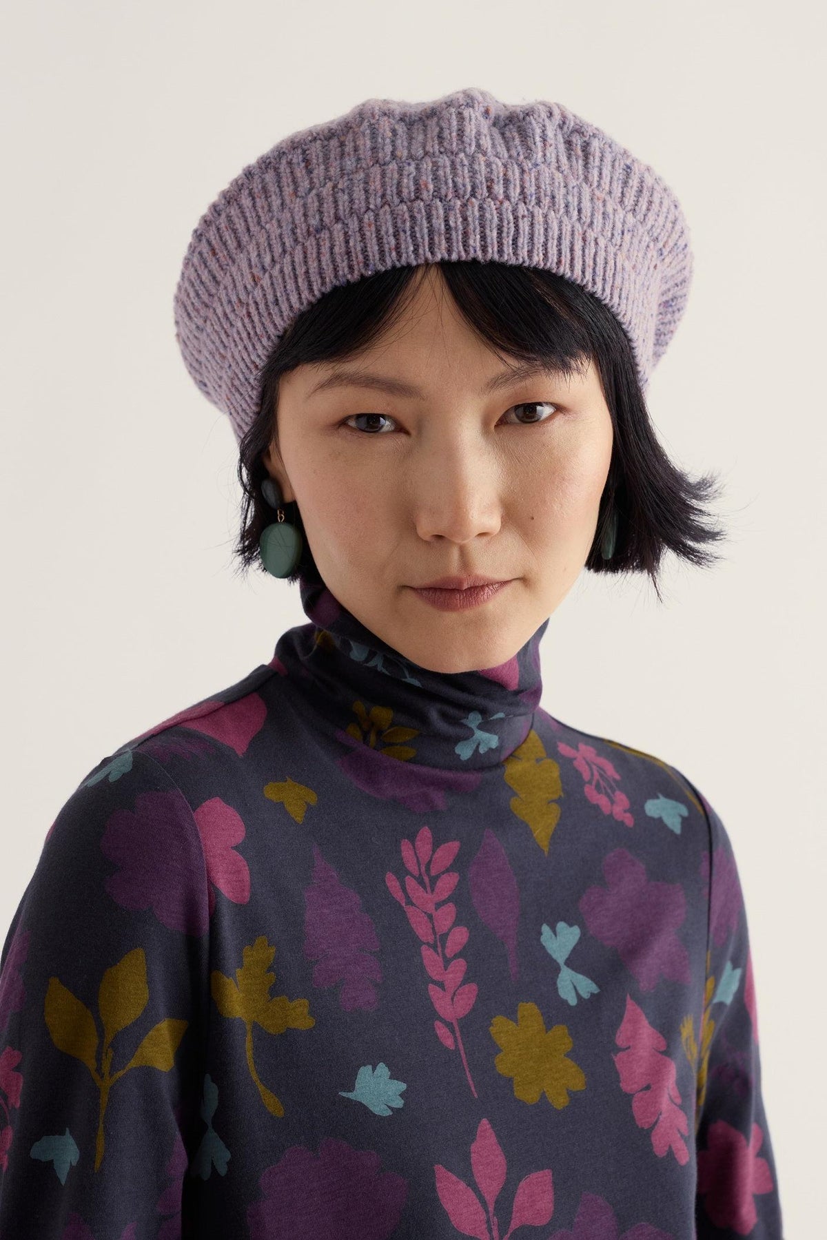 A woman wearing a textured purple merino wool beret and a dark floral print turtleneck top.