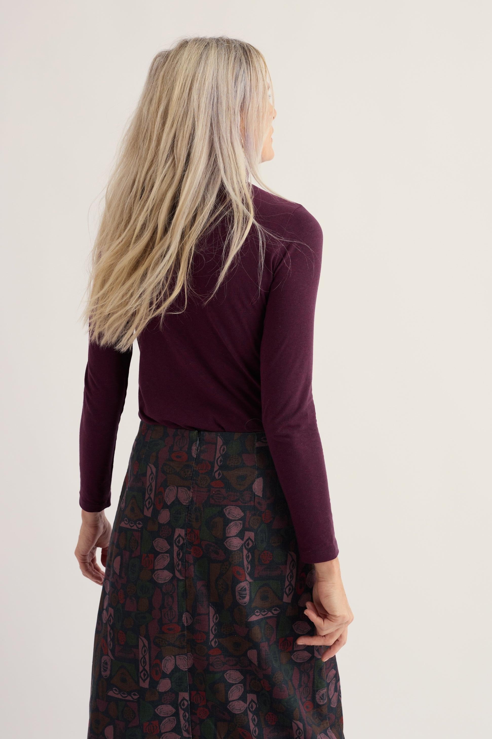 Woman seen from behind wearing a long-sleeved purple organic cotton roll neck top and a patterned skirt.
