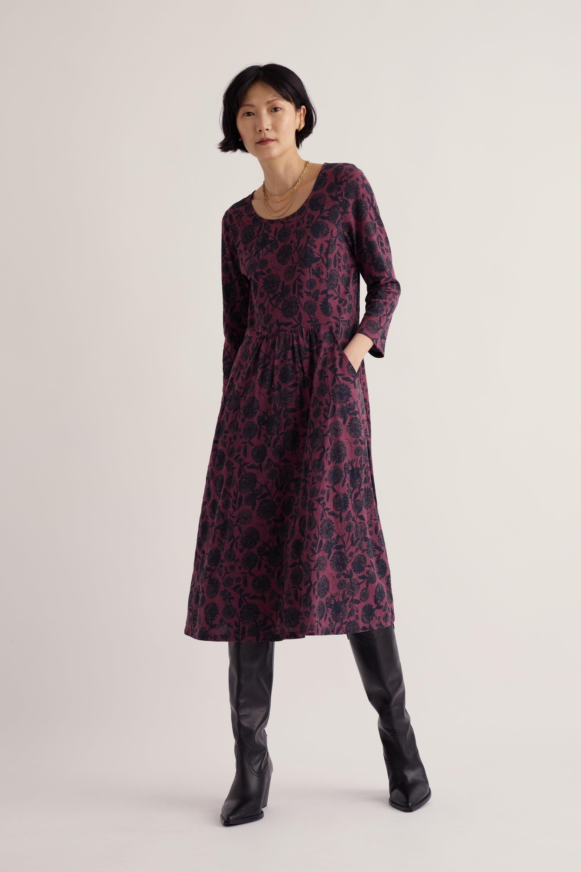 A woman posing in a purple knee-length Seasalt Cornwall casual dress with 3/4 length sleeves and black boots.