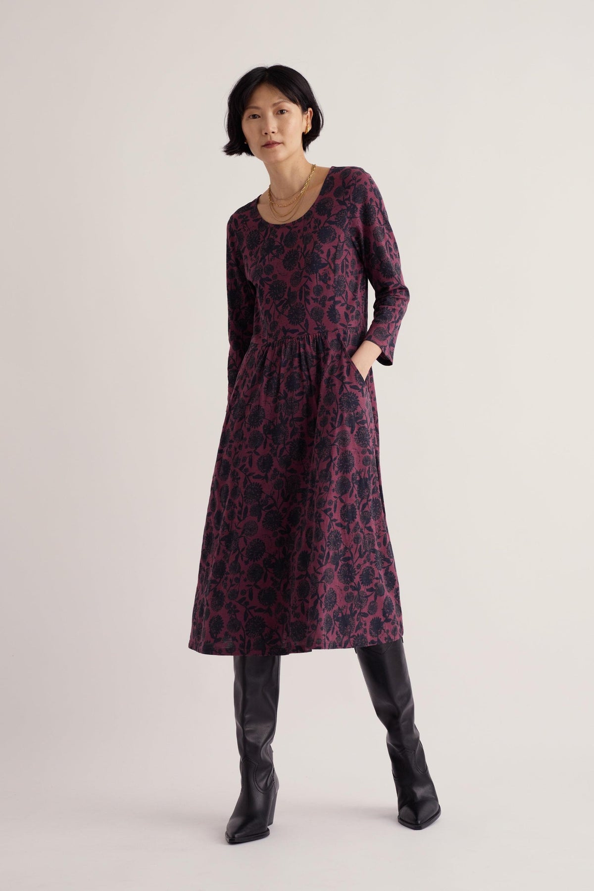 A woman posing in a purple knee-length Seasalt Cornwall casual dress with 3/4 length sleeves and black boots.