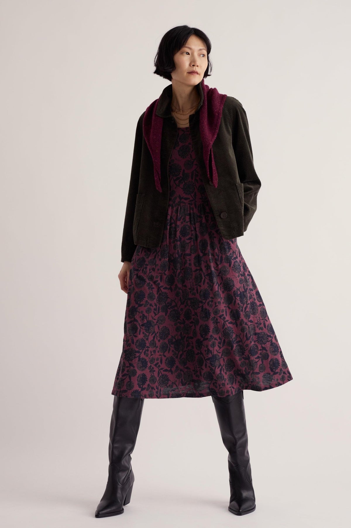 A woman posing in a purple floral Seasalt casual dress with a dark green corduroy jacket and black boots.