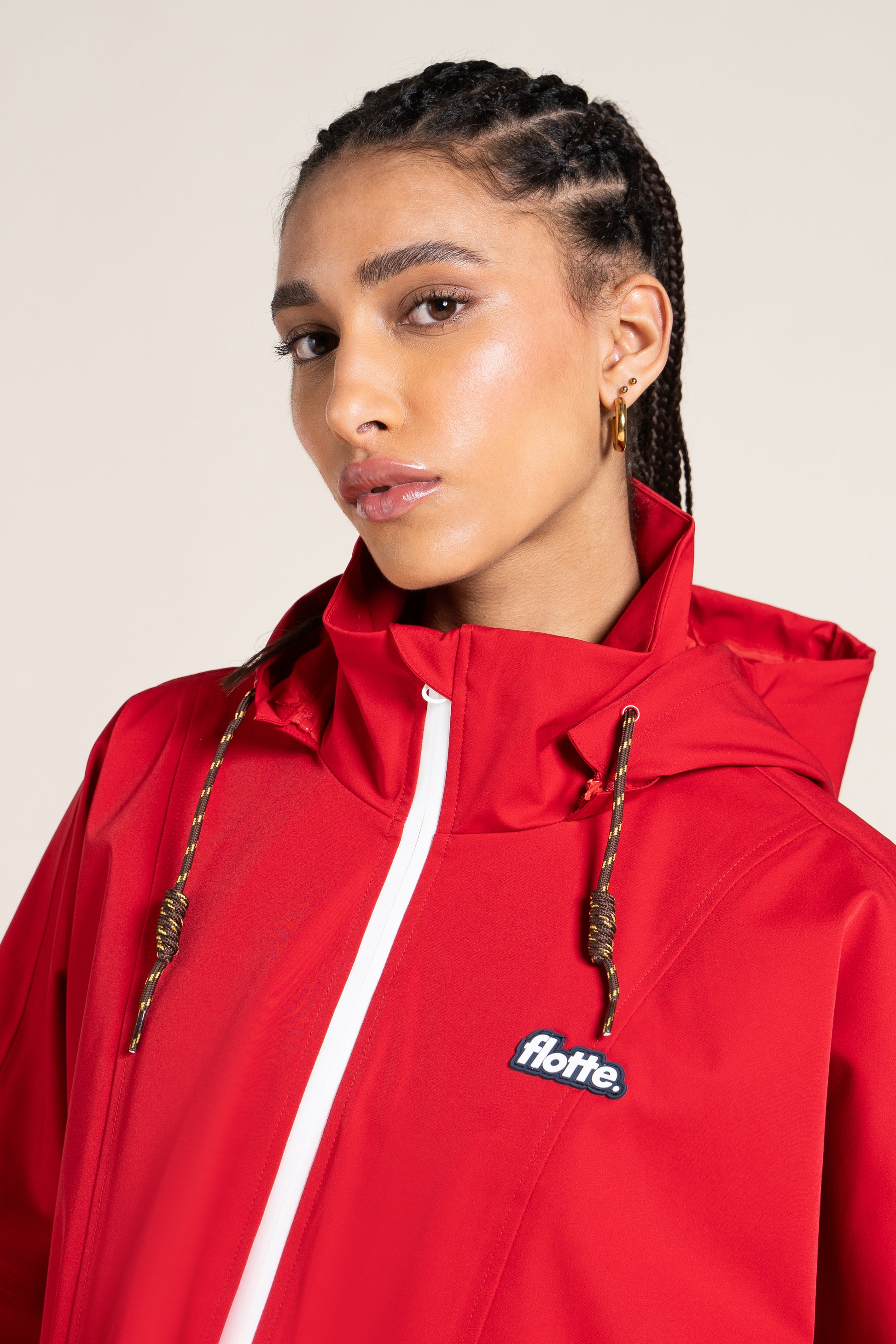 A woman with braided hair wearing a bright red Flotte branded rain jacket against a neutral studio background.