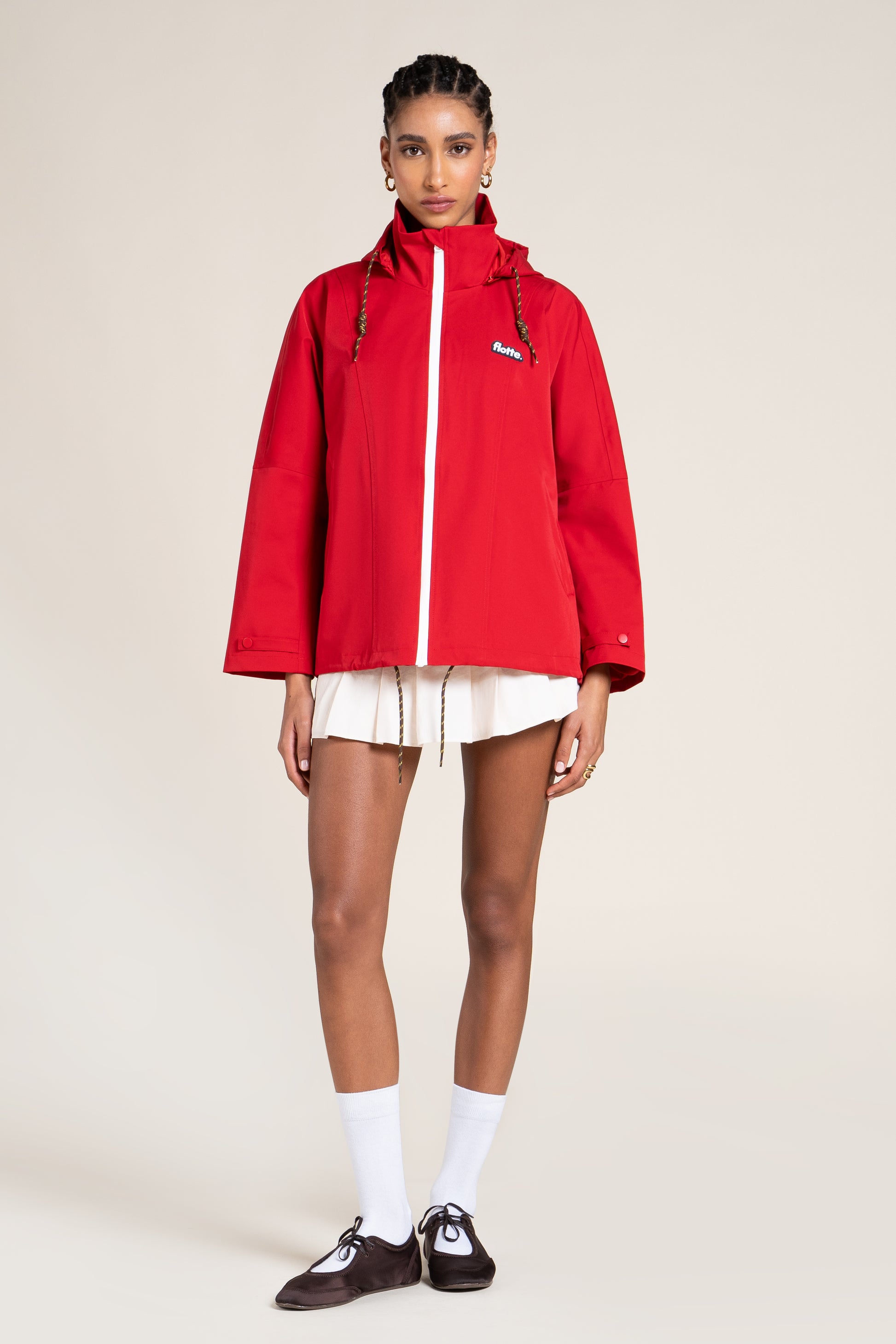 Model wearing a bright red Flotte raincoat with a white pleated skirt against a neutral studio background.