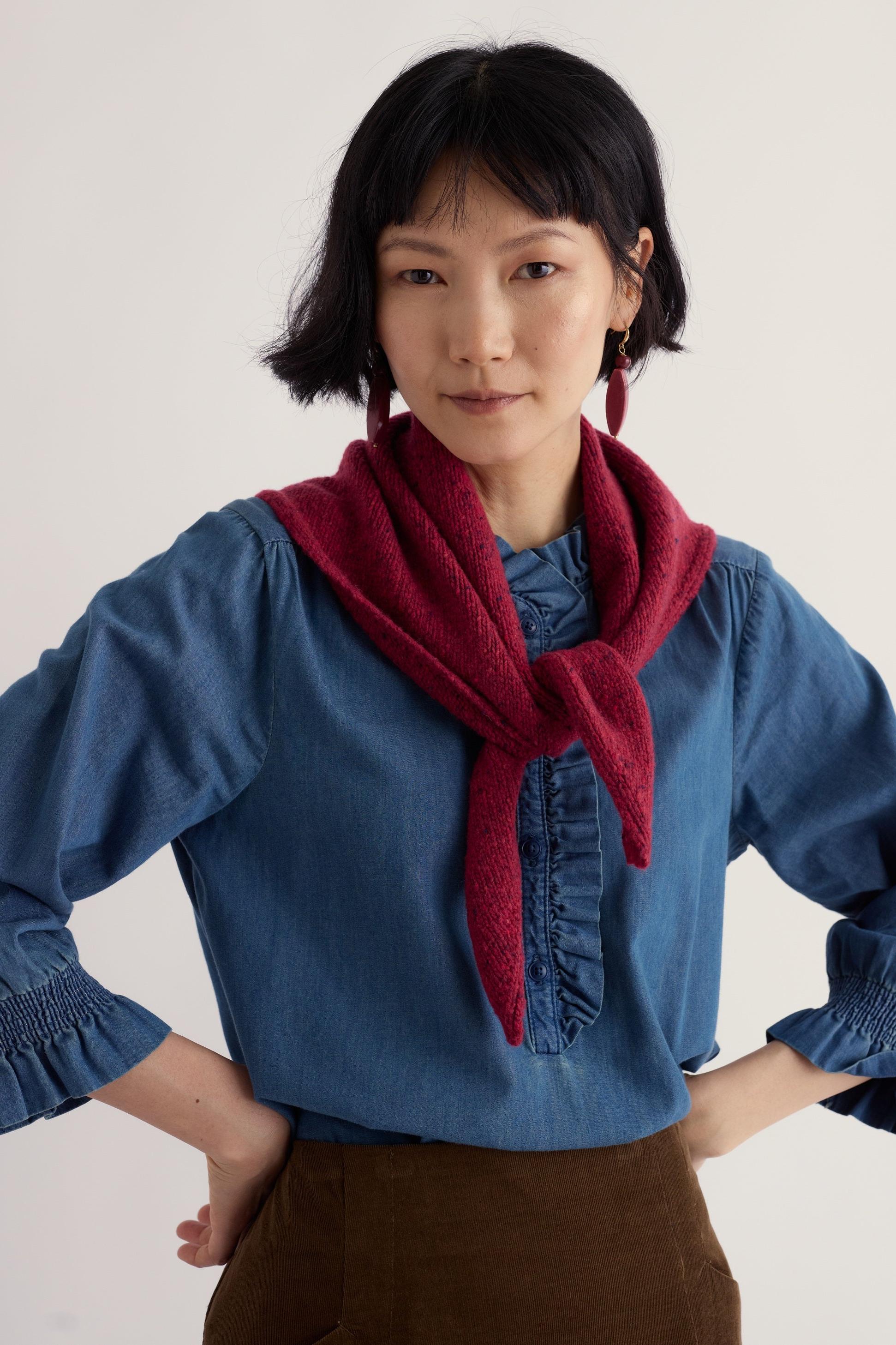 A woman wearing a vibrant red knit scarf over a blue ruffled denim shirt against a neutral studio background.