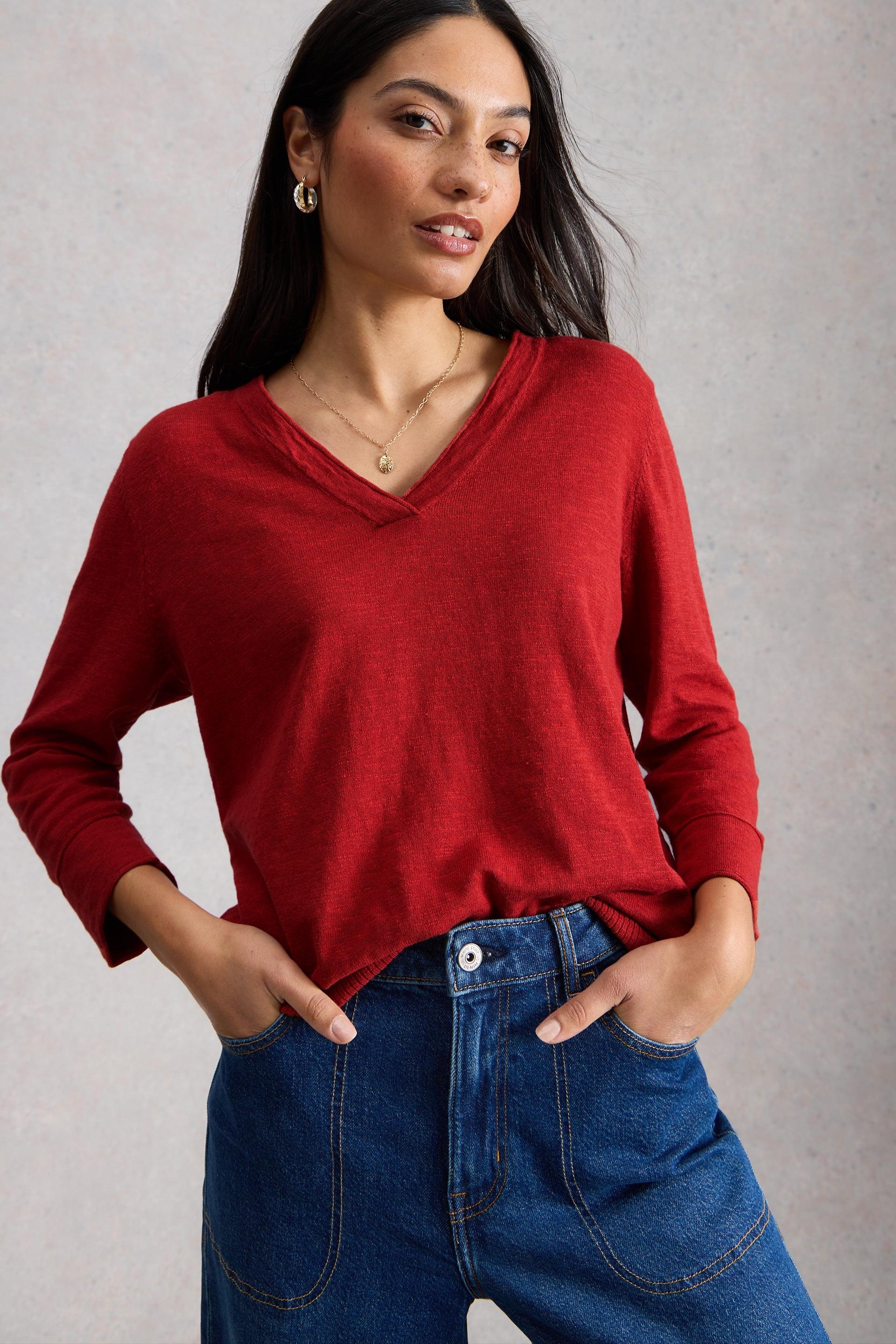 Woman wearing a red v-neck sweater made of organic cotton with her hands in her blue jeans pockets.