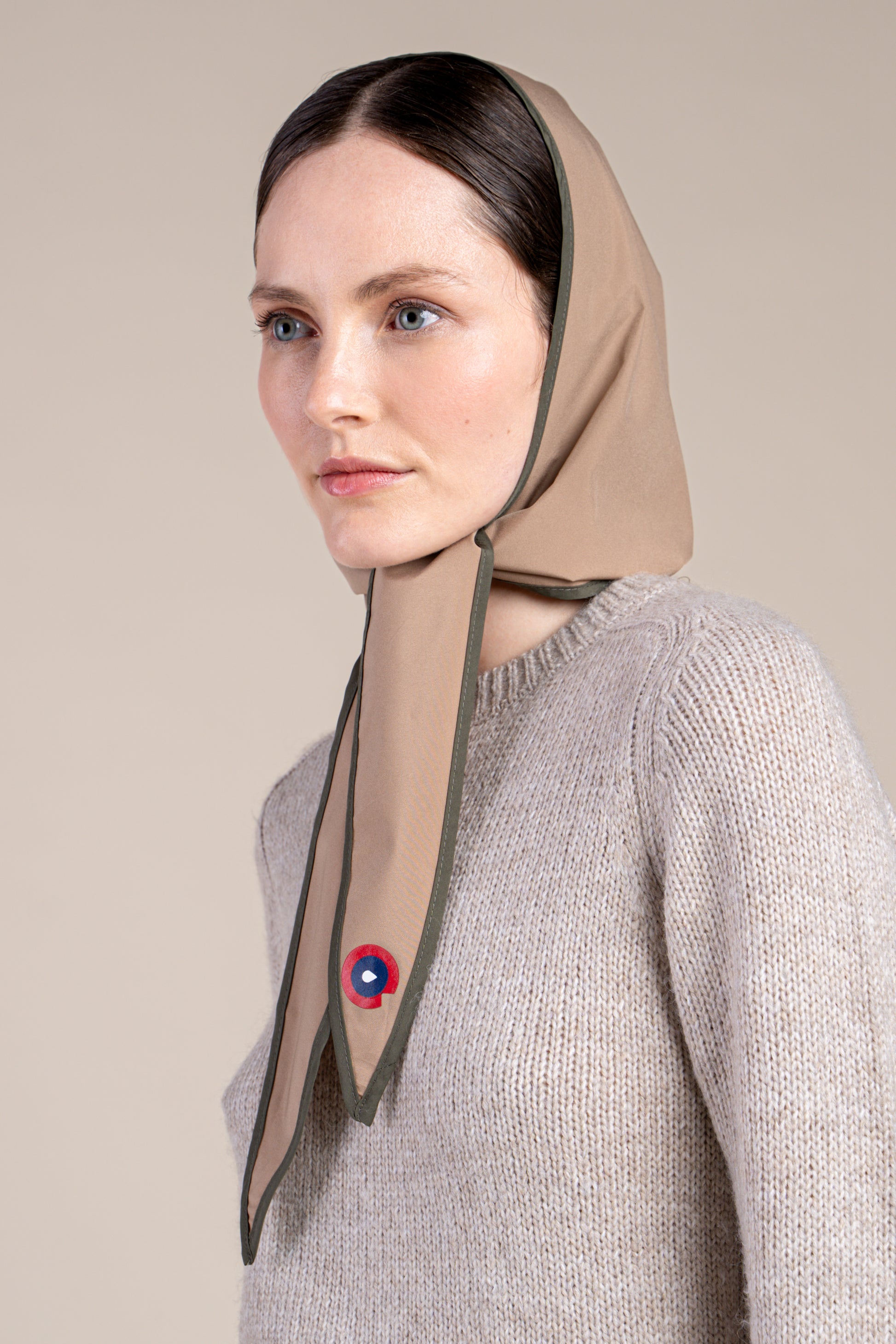Woman wearing a sahara colour waterproof head covering and a beige knit sweater against a neutral background.