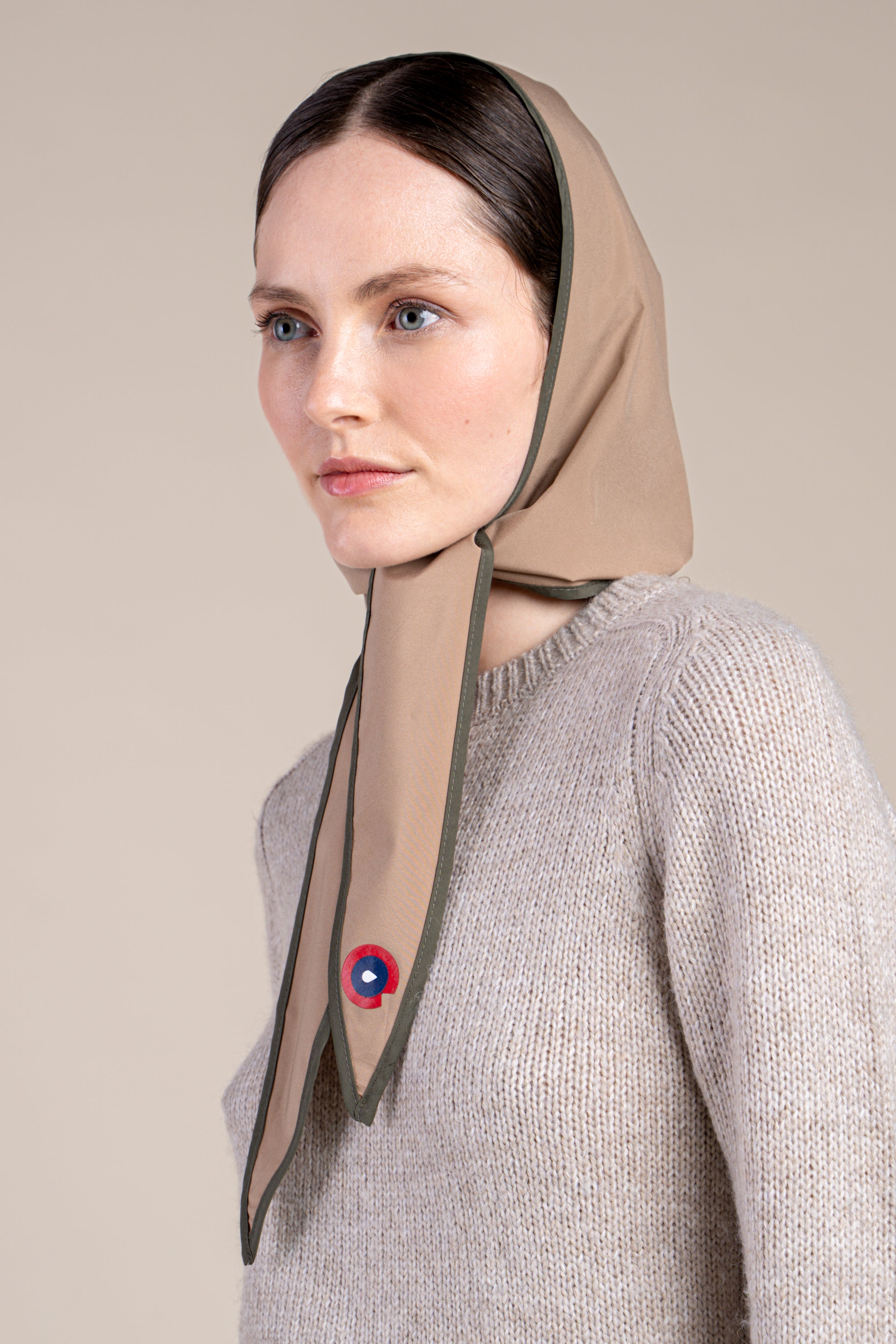 Woman wearing a sahara colour waterproof head covering and a beige knit sweater against a neutral background.