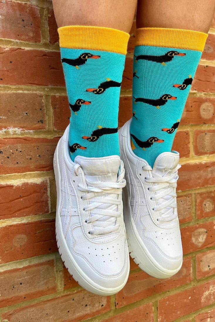 A person wearing turquoise bamboo sausage dog socks and white sneakers against a red brick wall background.