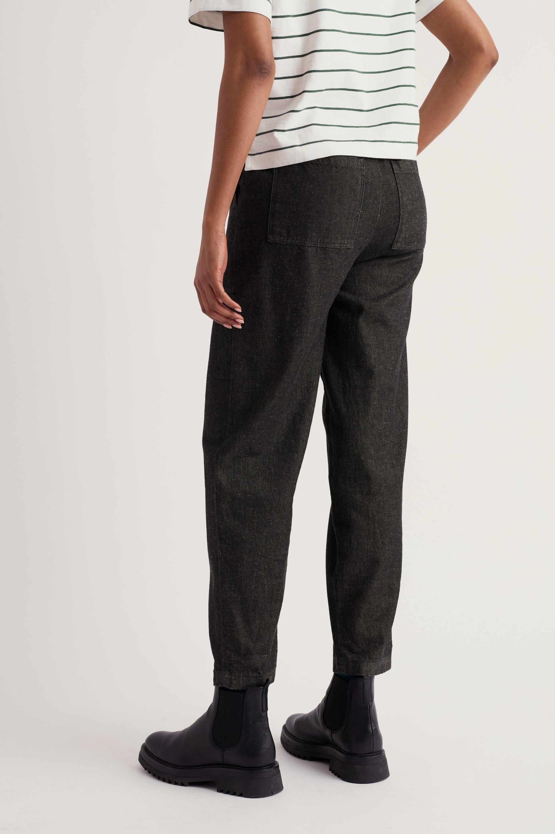 A person standing in casual black organic cotton trousers paired with black ankle boots against a white studio background.