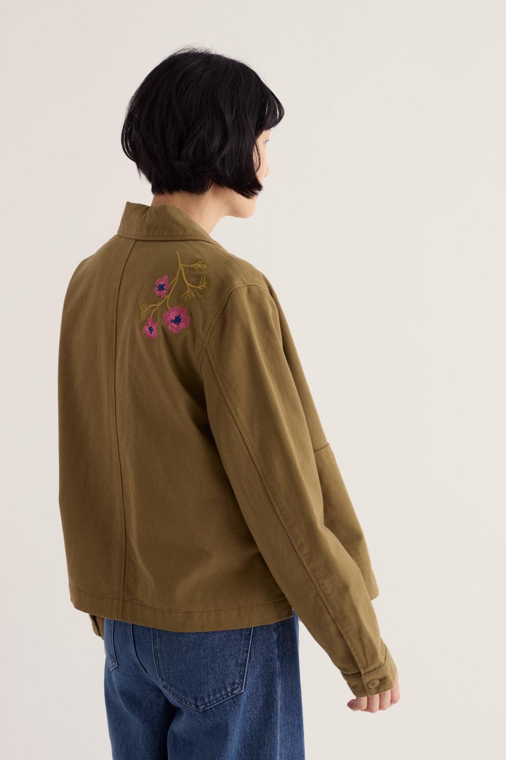 Woman seen from behind wearing an embroidered khaki Seasalt organic cotton jacket with blue jeans.