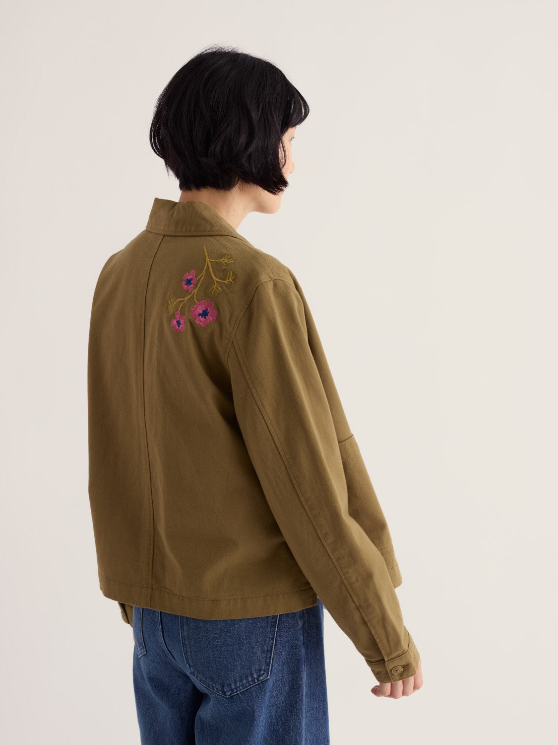 Woman seen from behind wearing an embroidered khaki Seasalt organic cotton jacket with blue jeans.