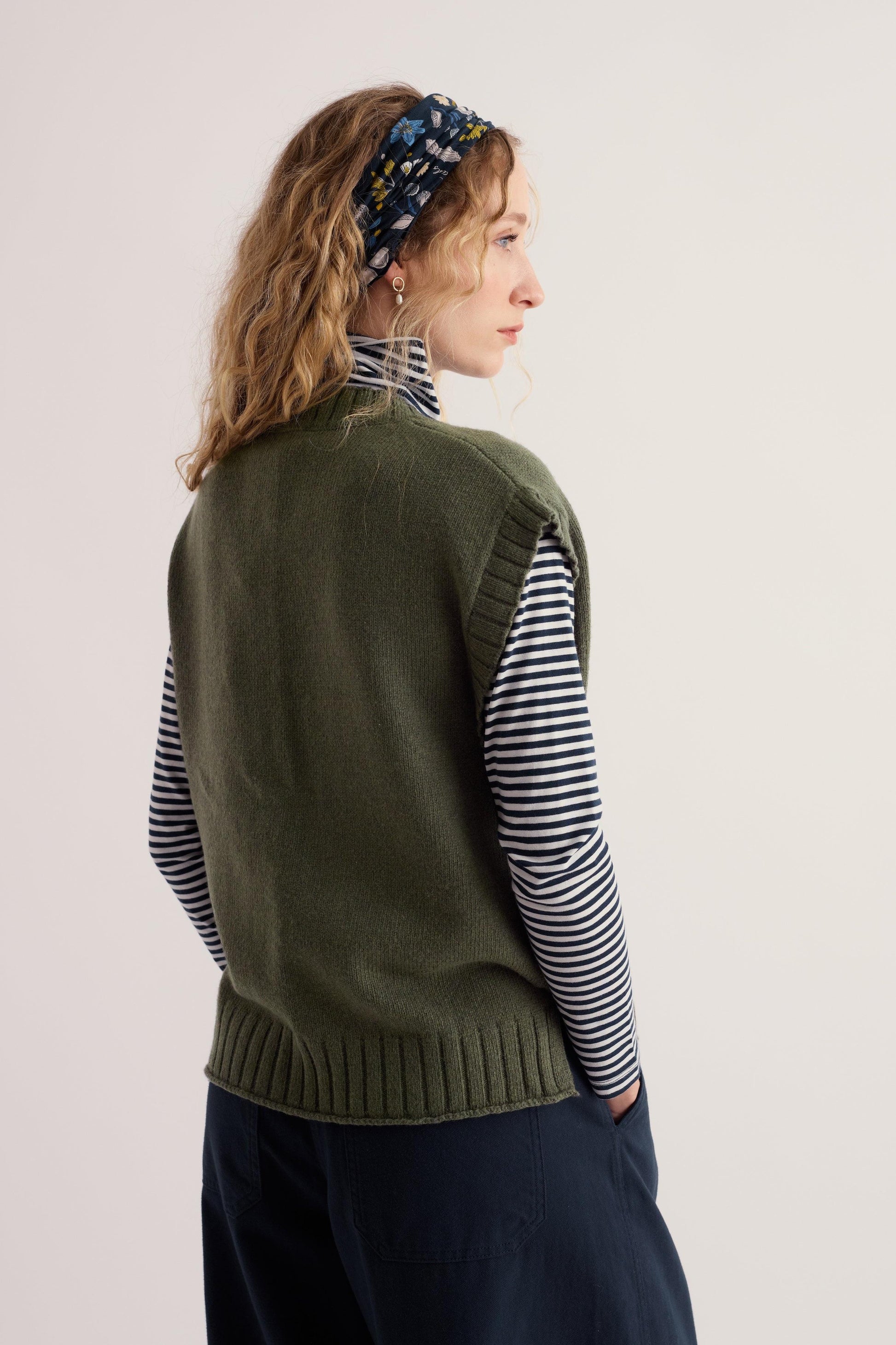 A woman wearing a khaki knit vest layered over a striped long-sleeve top against a neutral background.