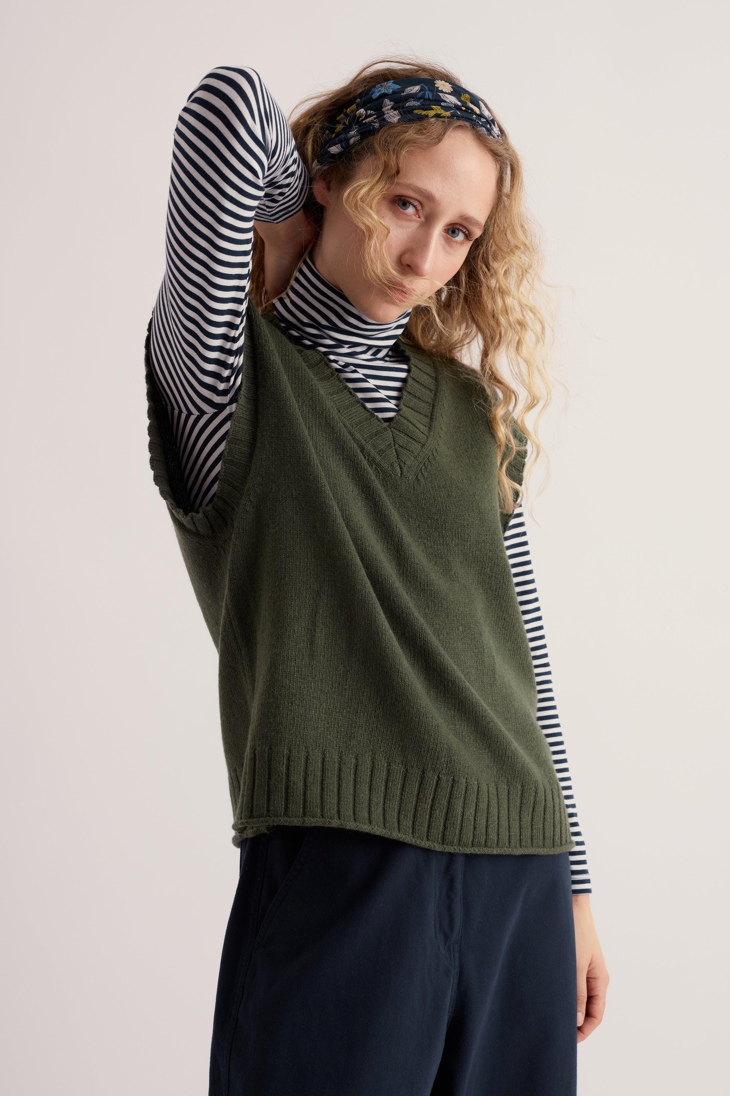 A woman wearing a khaki knit vest layered over a striped long-sleeve top against a plain background.