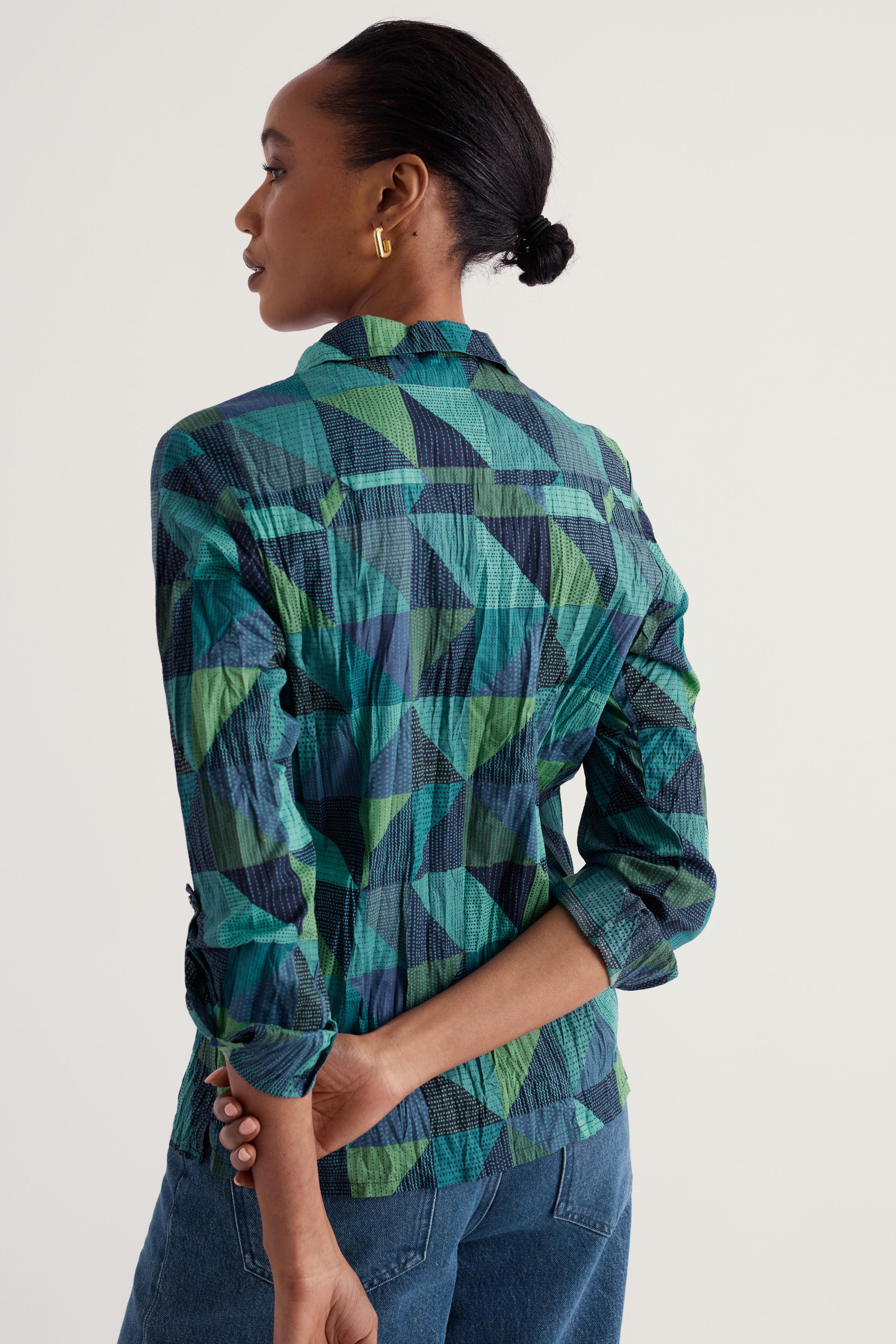 A woman shown from the back wearing a long-sleeve button-up shirt with a green, teal, and navy geometric pattern, paired with blue jeans against a plain light background.
