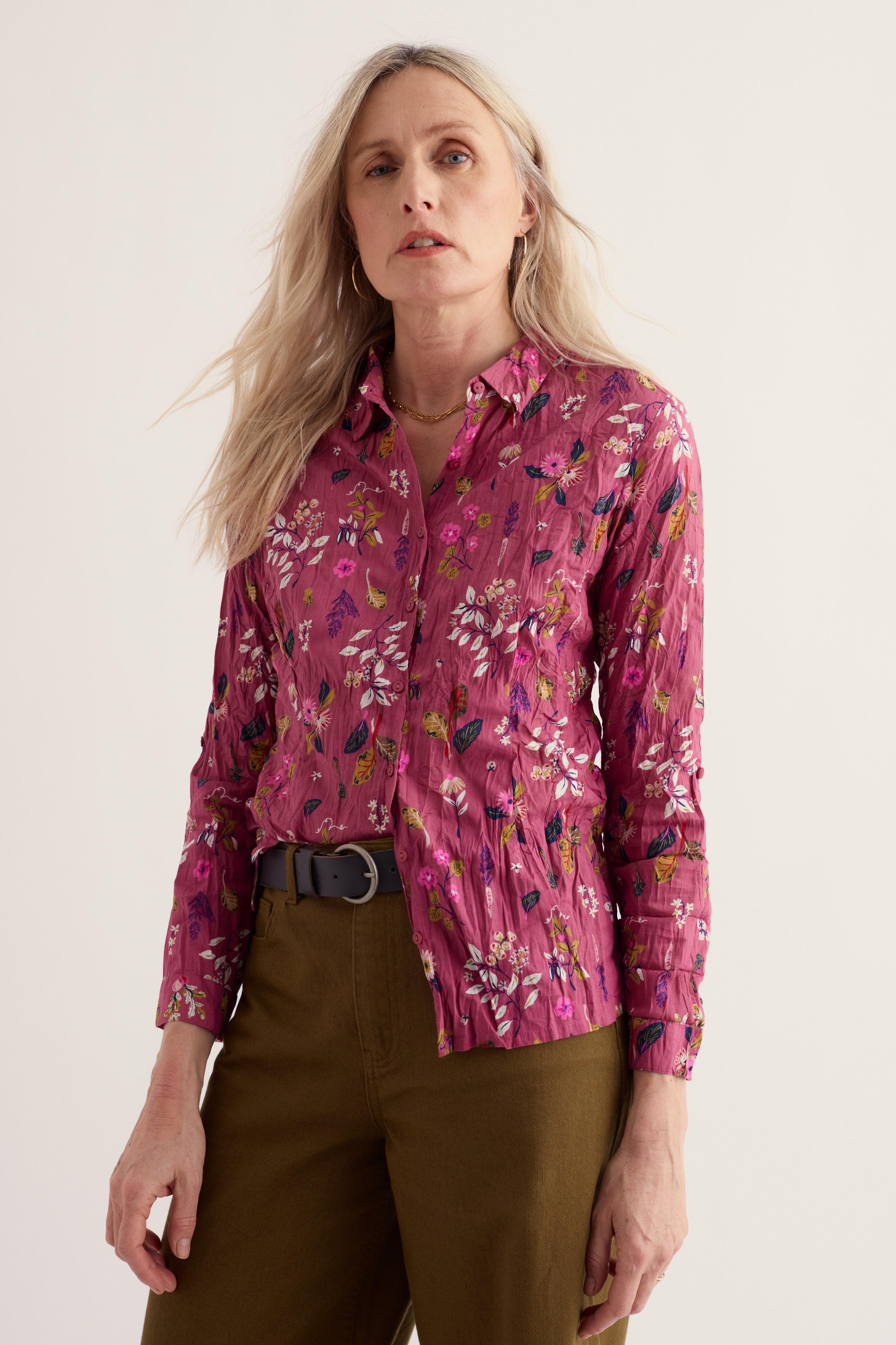 A woman wearing a long-sleeve button-up shirt with a pink and purple floral pattern, paired with olive green trousers, photographed against a plain light background.