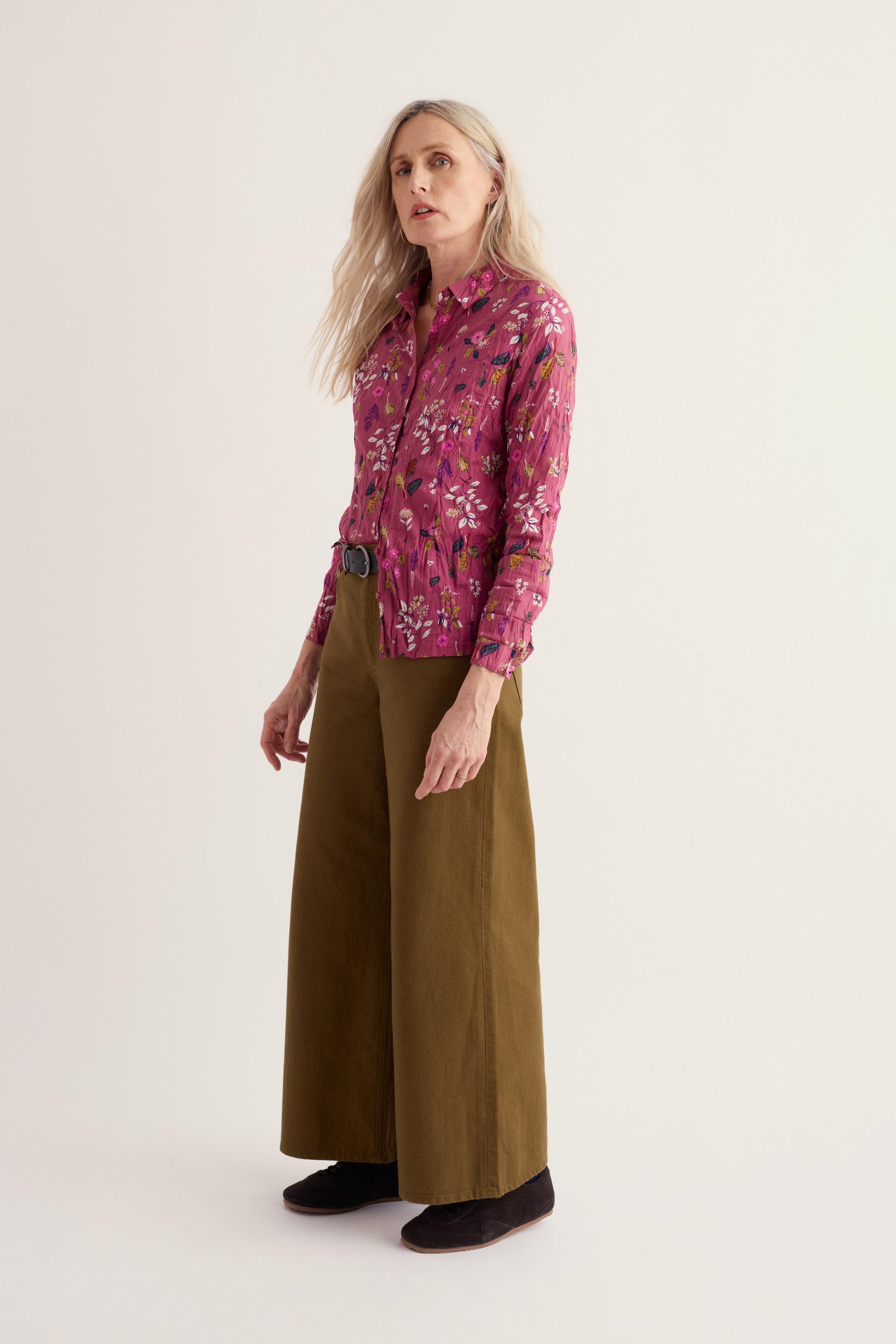 A woman modeling a long-sleeve button-up shirt with a pink and purple floral pattern, paired with wide-leg olive green trousers and black shoes, against a plain light beige studio background.