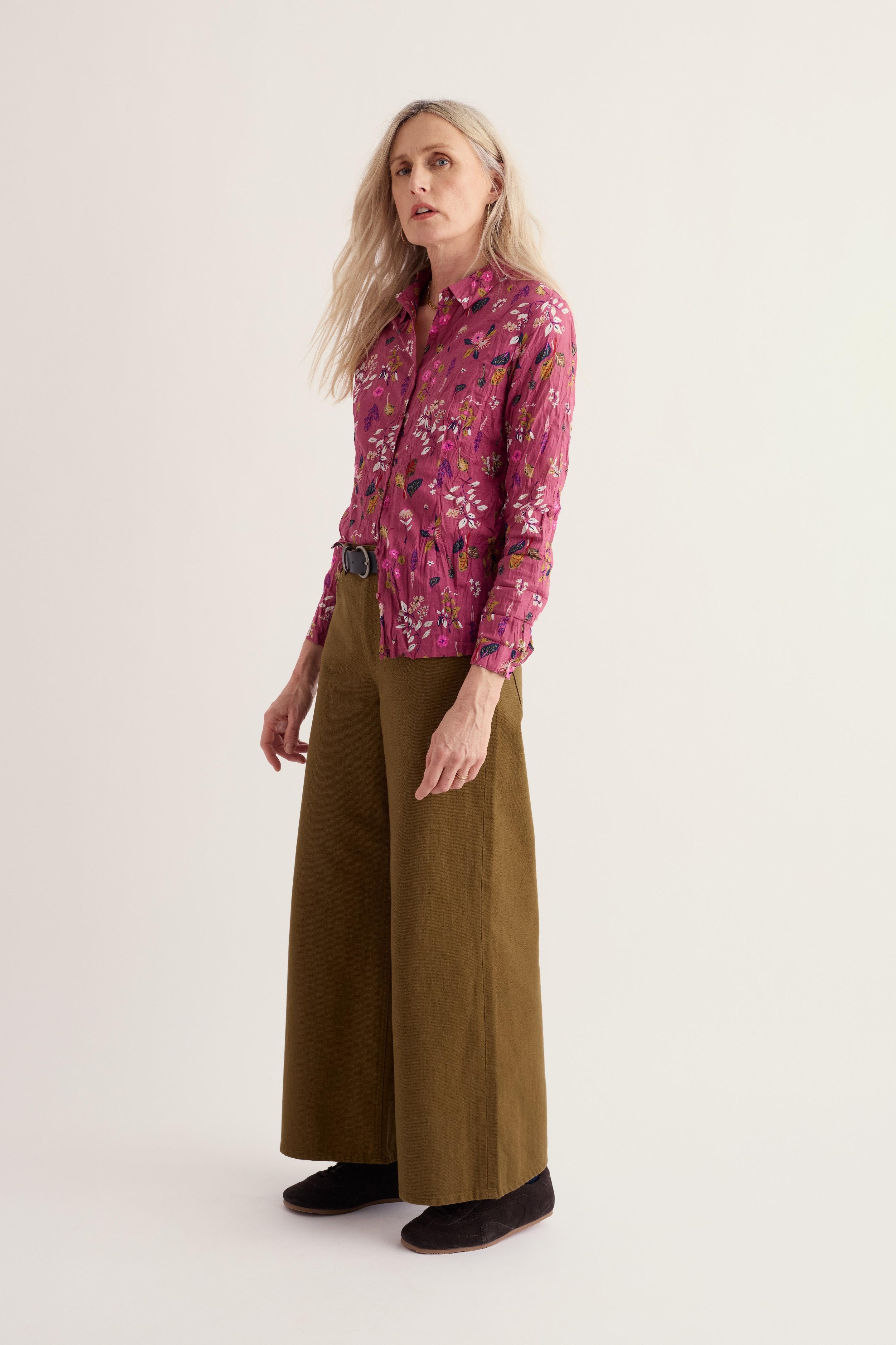A woman modeling a long-sleeve button-up shirt with a pink and purple floral pattern, paired with wide-leg olive green trousers and black shoes, against a plain light beige studio background.