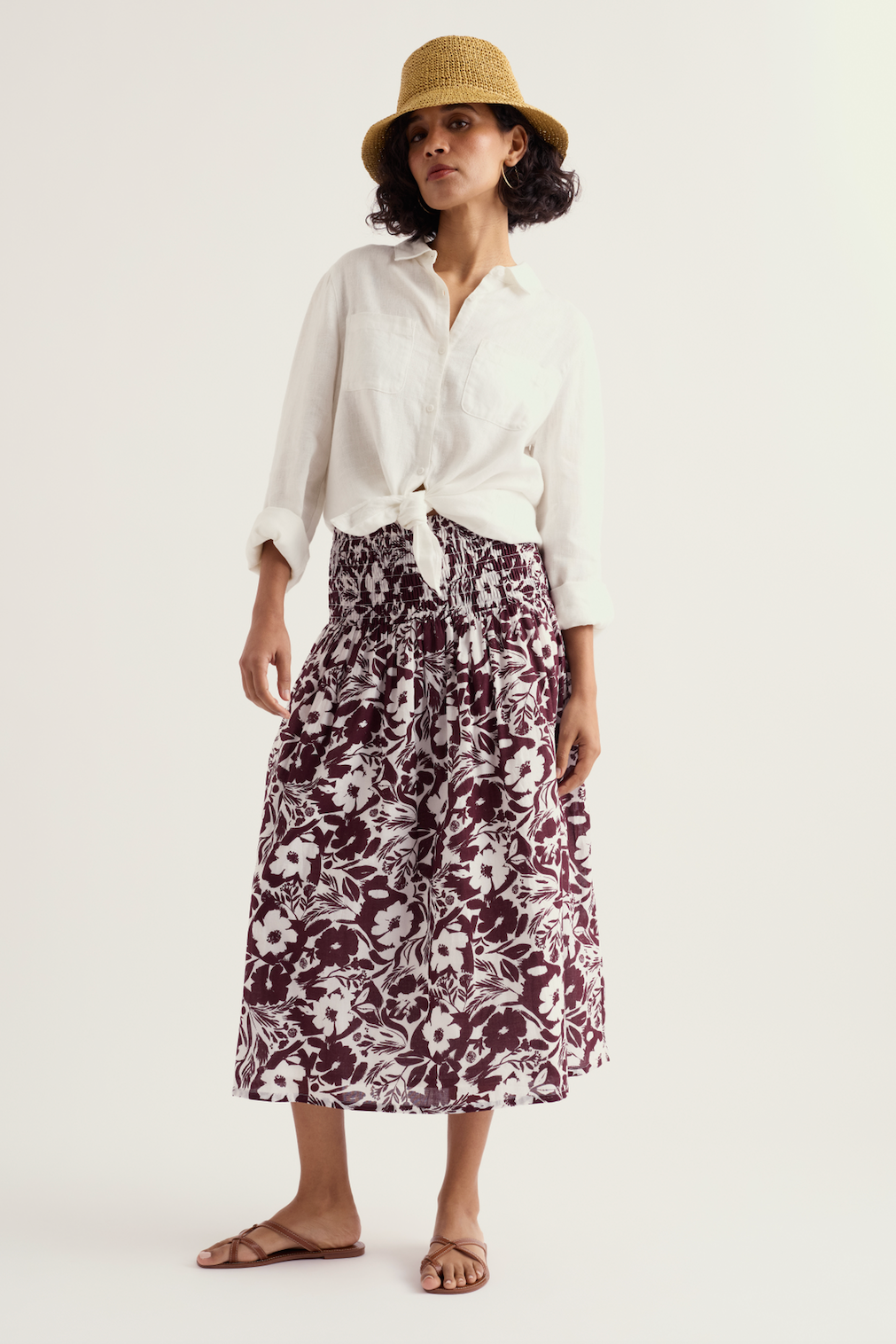 Woman wearing a floral print Seasalt organic cotton skirt and white button-down shirt with a straw hat.