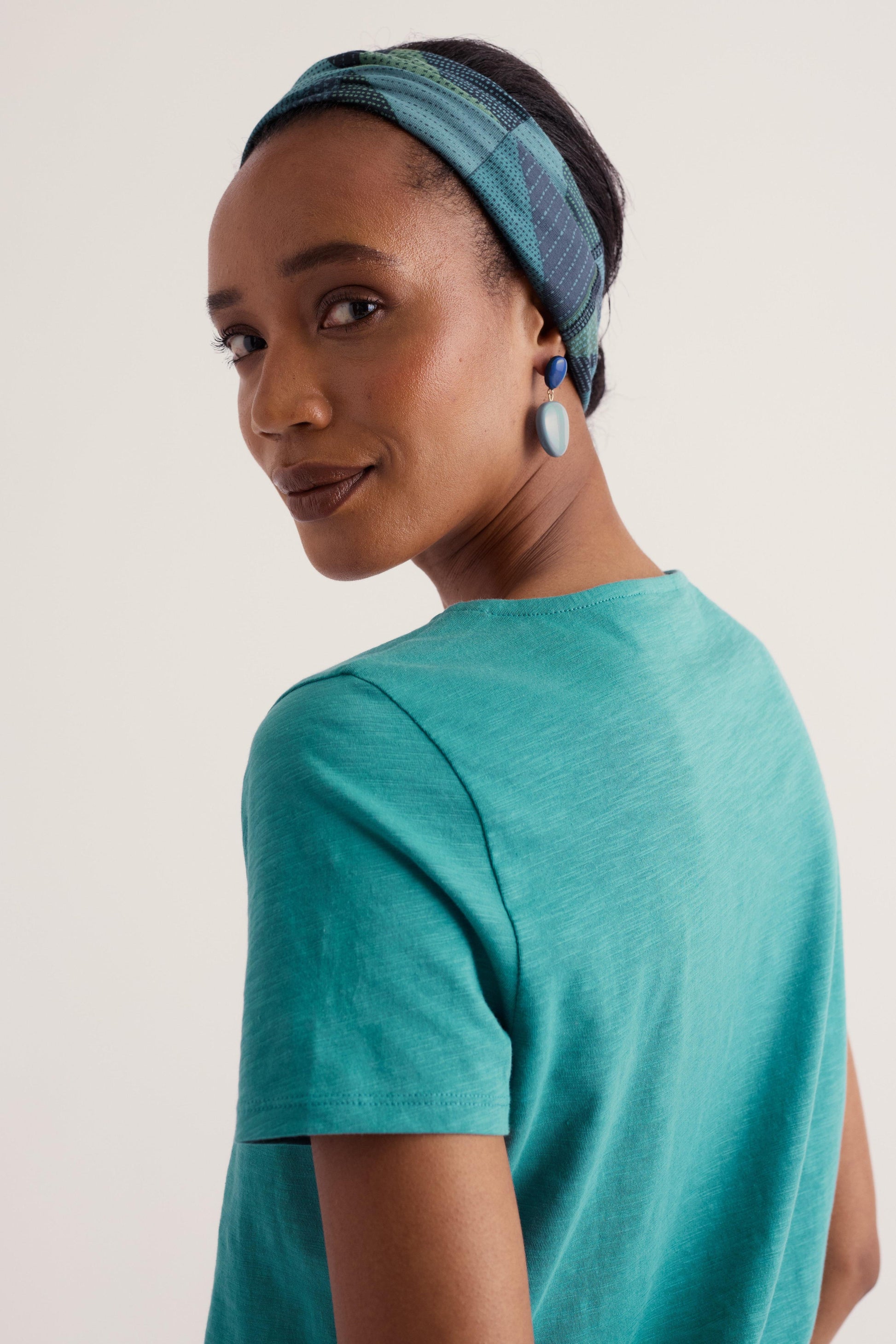 A woman wearing a teal short-sleeve t-shirt and a blue patterned headband, shown from a side-back angle against a plain light background.