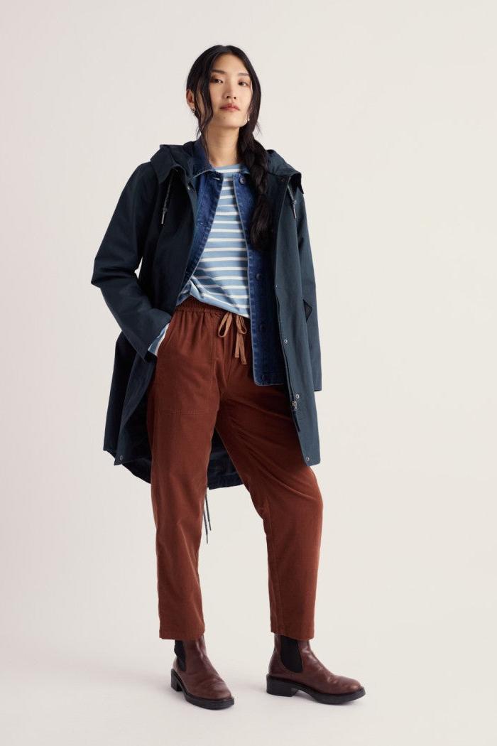 A model stands against a plain light beige studio background wearing a dark navy hooded jacket over a blue and white striped shirt, paired with rust-brown drawstring trousers and dark brown Chelsea boots.