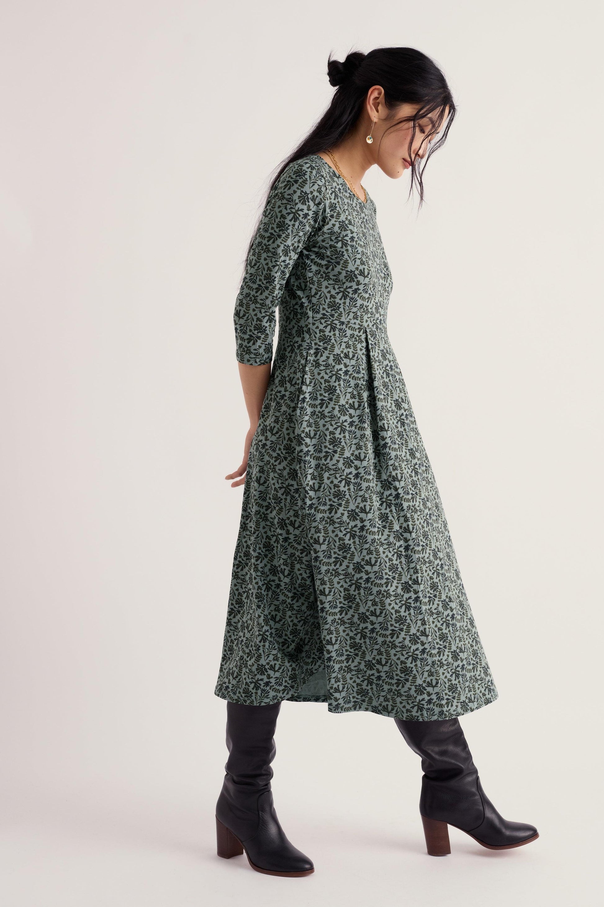 A woman wearing a green and white floral print midi dress with three-quarter sleeves, paired with black knee-high leather boots, photographed against a plain light background.