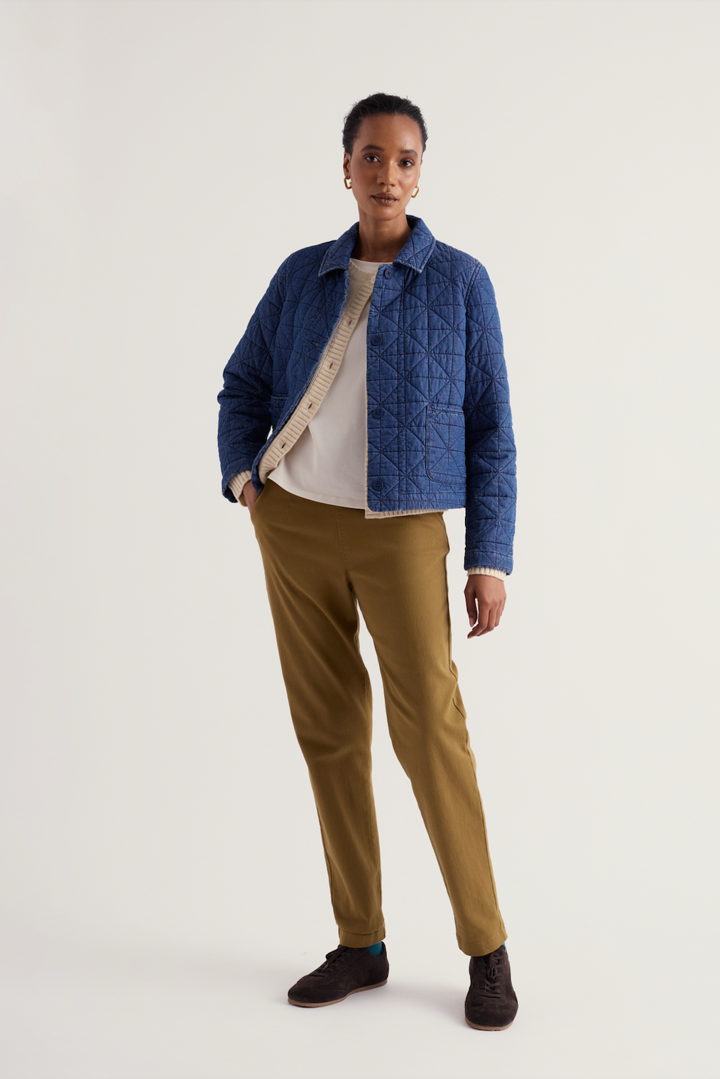 A model stands against a light gray studio background wearing a blue quilted jacket over a cream knit top, olive-brown trousers, and dark brown shoes.