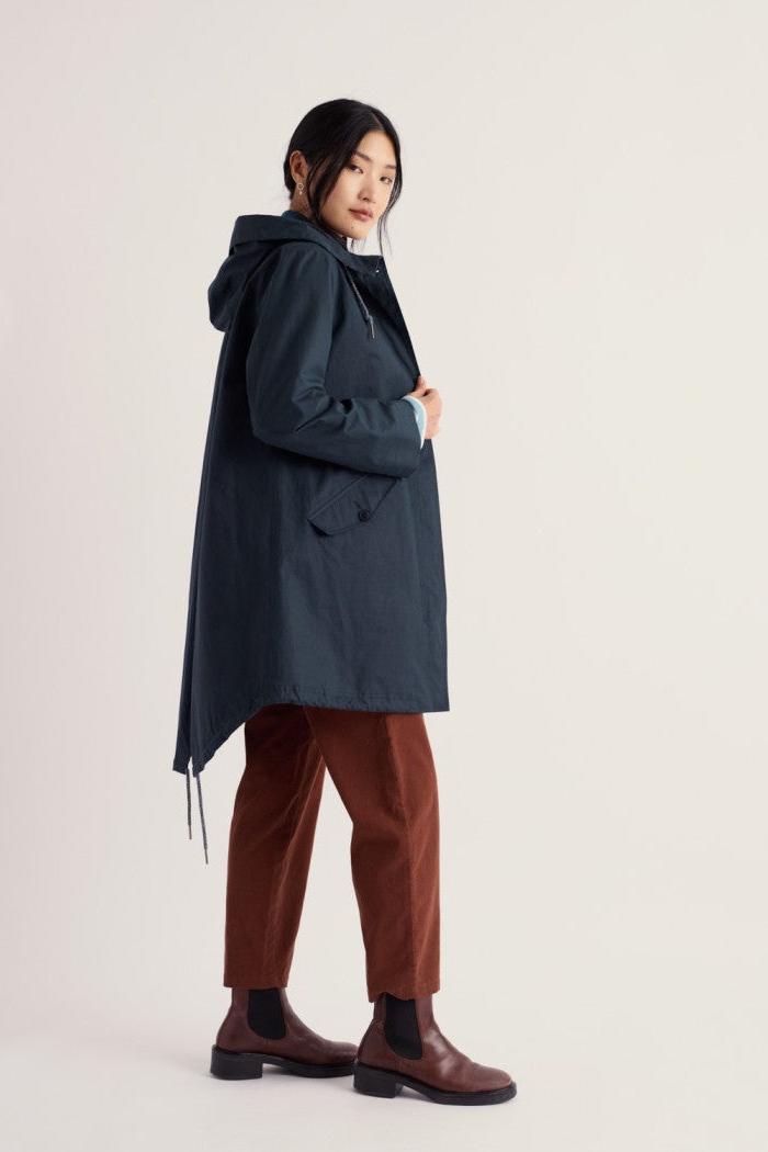 A woman modeling a long hooded coat in a dark slate blue color, paired with rust-brown trousers and brown Chelsea boots, against a plain light beige studio background.