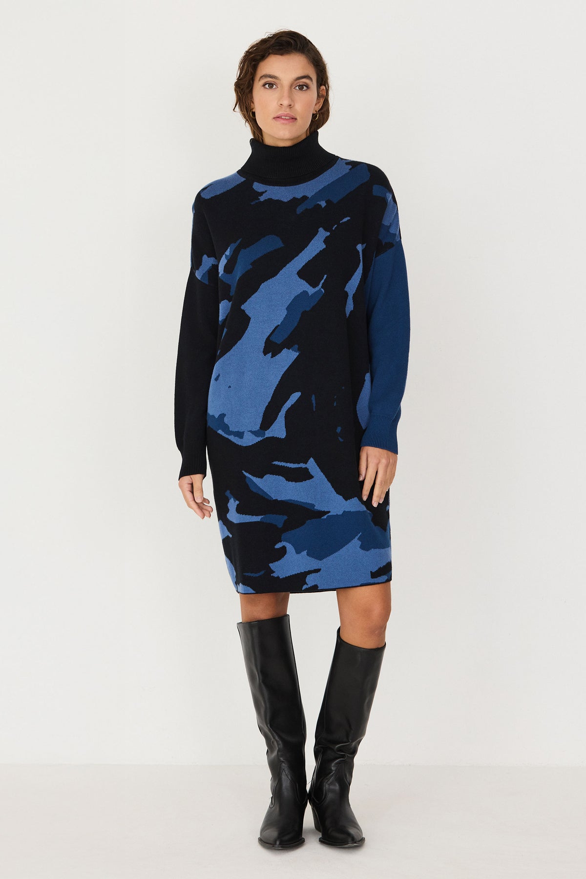 Woman standing in a black and blue SKFK cotton knit turtleneck dress paired with black boots.