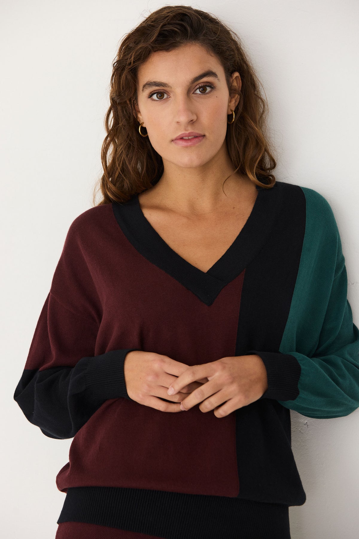 A woman with wavy brown hair wearing a colour-blocked V-neck cotton jumper by SKFK against a white wall.