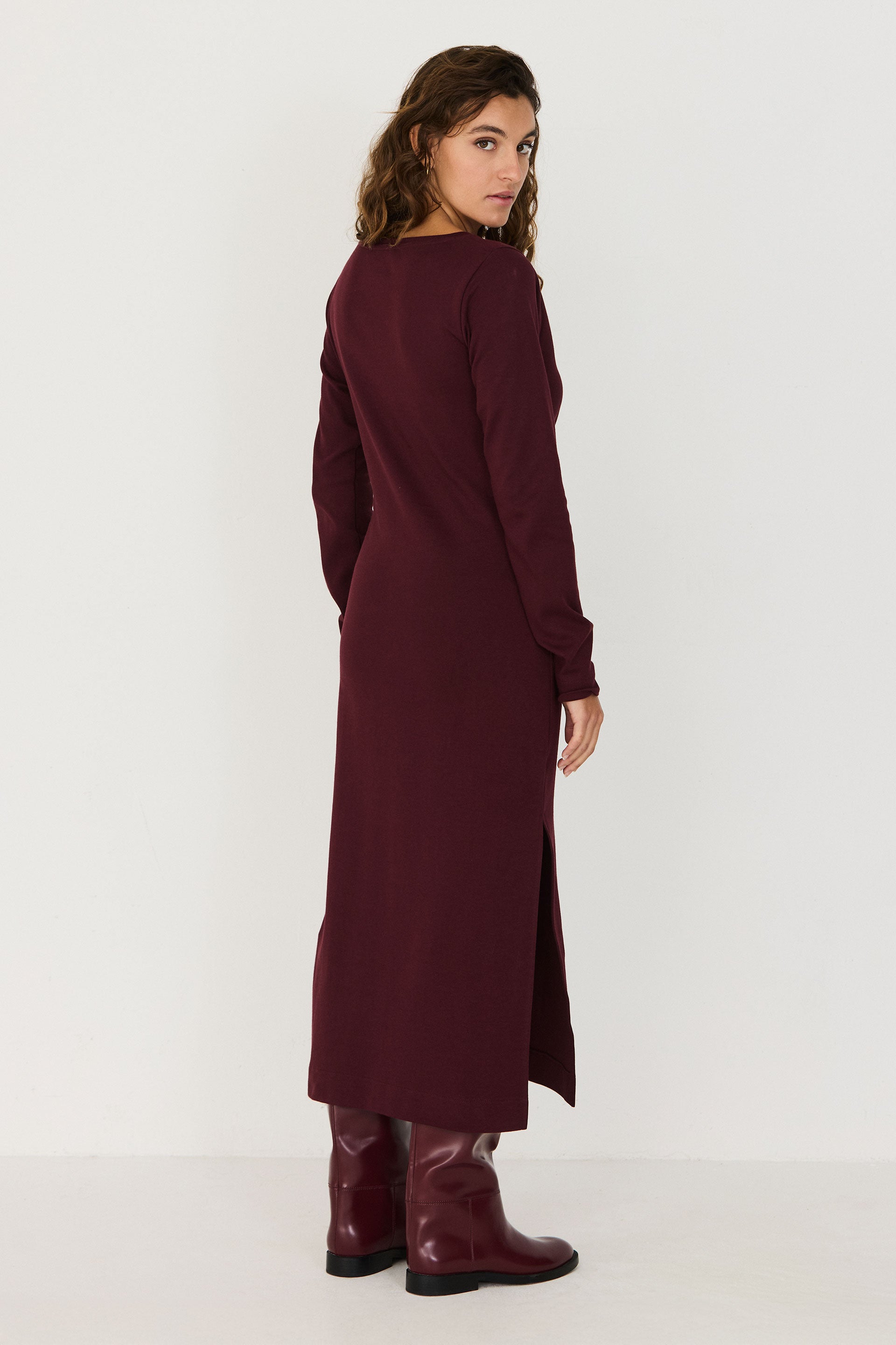 A woman wearing a chocolate brown long-sleeved organic cotton midi dress by SKFK standing against a white wall.
