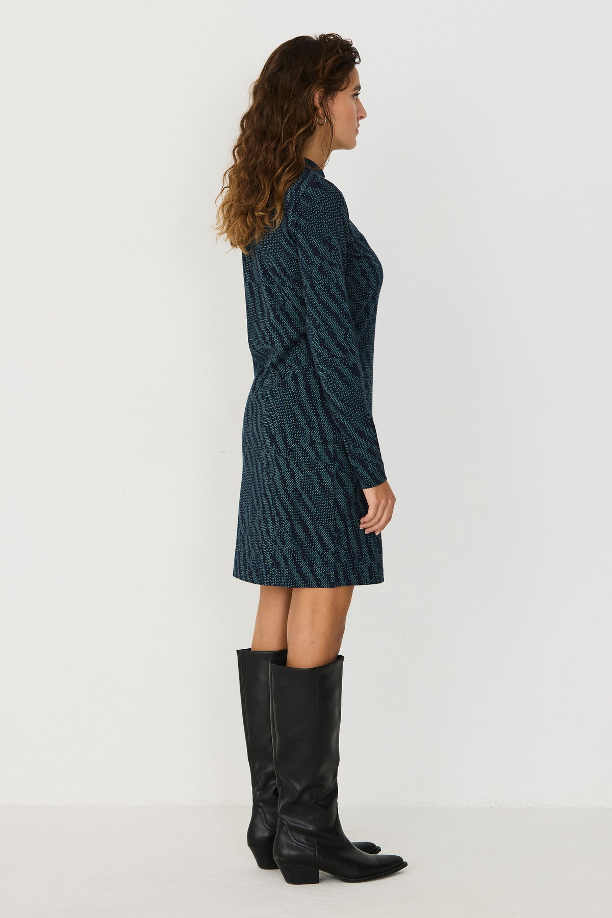 Profile view of a woman wearing a SKFK patterned green casual dress and black knee-high boots against a white background.